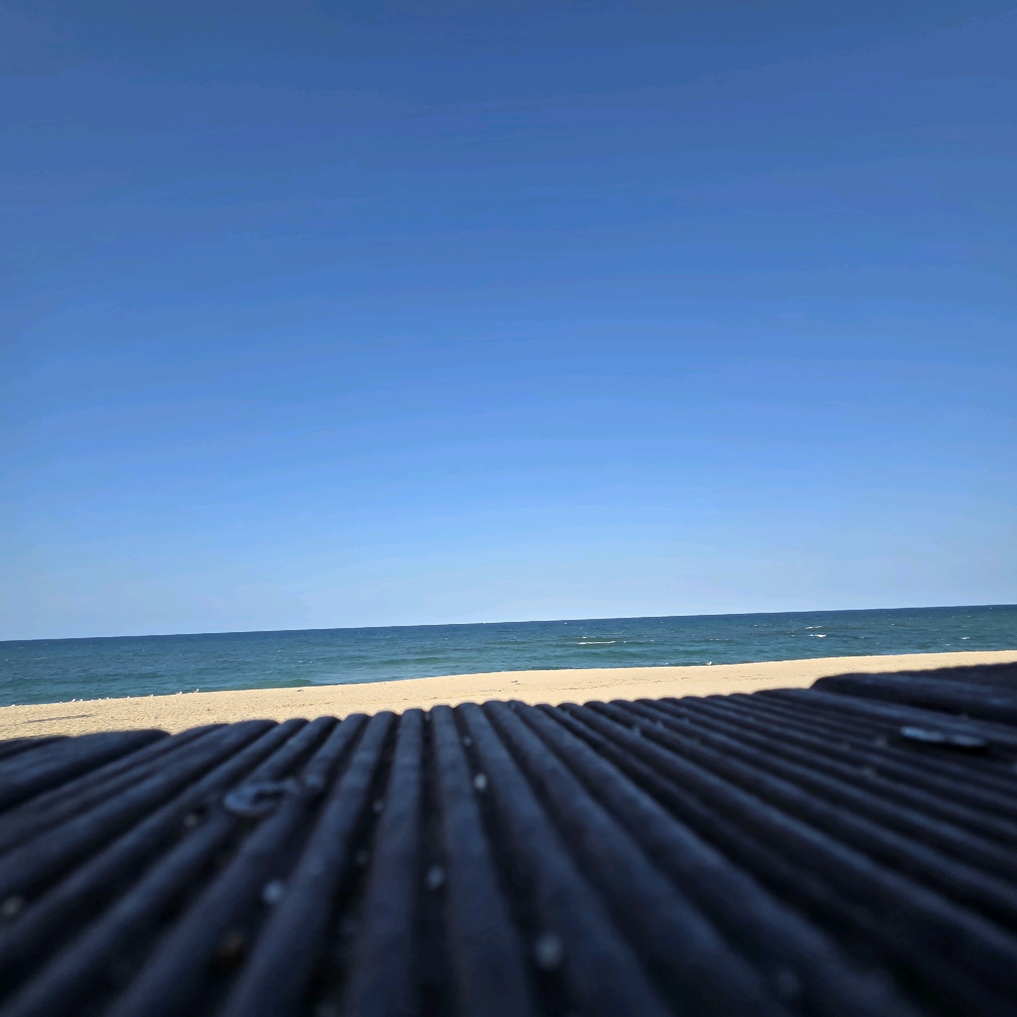 This is a photograph of a beach scene in Gangneung-si, South Korea. The image is taken from a low angle, looking out over a dark, ribbed surface that appears to be a bench or boardwalk railing in the foreground. Beyond the railing, a sandy beach stretches towards the ocean. The water is a deep blue with whitecaps, indicating waves. The horizon line is sharp and clear against a bright, cloudless blue sky. A thin strip of sand is visible between the dark foreground element and the water. There are a few small white objects, possibly seagulls, on the beach in the distance. The overall impression is one of a calm, sunny day at the seaside.