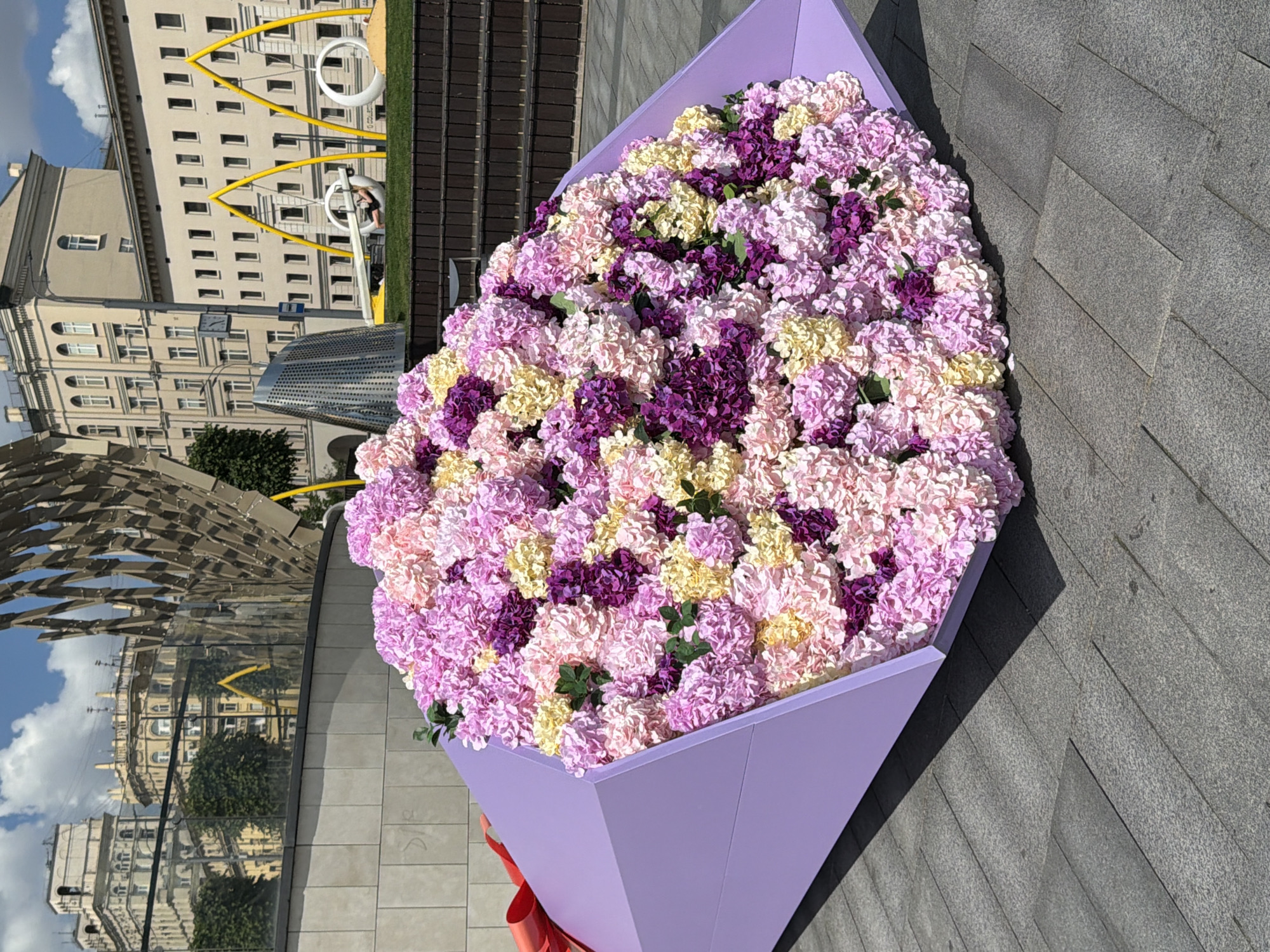 This image, captured in Moscow, Russia, showcases a large, vibrant public art installation resembling an oversized flower bouquet in an urban setting. The main subject is a giant arrangement of artificial flowers, primarily hydrangeas or similar clusters, in striking shades of light pink, deep purple, and pale yellow. These flowers are presented in a massive, light purple, upward-tapering holder, designed like a gift box or floral wrap, with a small red ribbon tied at its bottom.
The installation rests on a paved area composed of grey, rectangular stone slabs, with clear shadows indicating bright sunlight, suggesting a sunny midday or afternoon. In the background, an assortment of multi-story buildings, mostly light-colored with numerous windows, defines the urban landscape. To the left, a modern, reflective architectural structure with a textured, possibly metallic or mirrored surface, reflects the sky and surrounding buildings. Yellow, curved artistic structures, potentially public art or recreational equipment, are visible behind the flower display. One cropped view shows a person interacting with a white circular element within these yellow structures. The sky above is clear with scattered white clouds. The scene conveys a bright, lively atmosphere in a contemporary urban environment. No discernible text is visible.