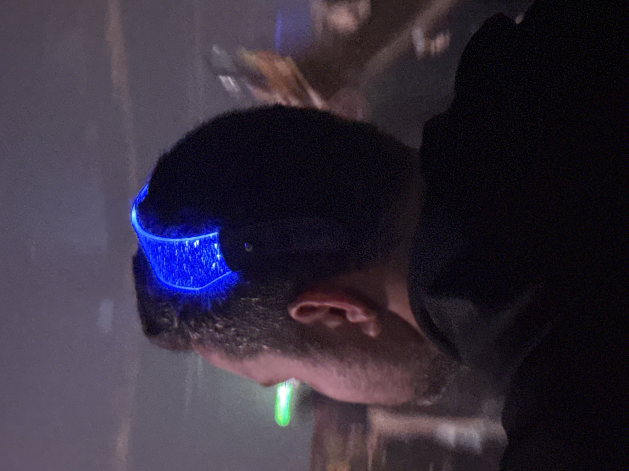 The image shows a close-up of a person wearing a blue, illuminated headband. The person has short, dark hair and a beard. The background is blurry and dark. There are some faint shapes and lines suggesting a room or building, but details are unclear. There is no indication of the location in the city of Khimki, Russia.