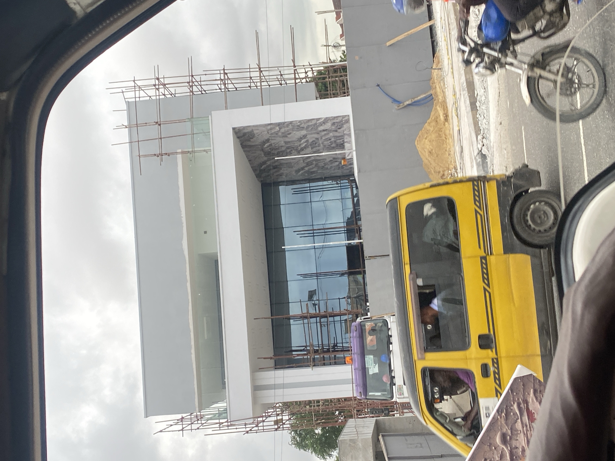 The image depicts a street scene in Ikeja, Nigeria, with a focus on a partially constructed modern building. Scaffolding surrounds the upper floors of the building, which features large glass windows and a grey facade with marble-patterned accents on the lower section. In the foreground, a yellow mini-bus, likely a public transport vehicle, is visible. To the right, a person is riding a motorcycle. The background shows more construction barriers and a pile of sand, indicating ongoing development. The sky is overcast.