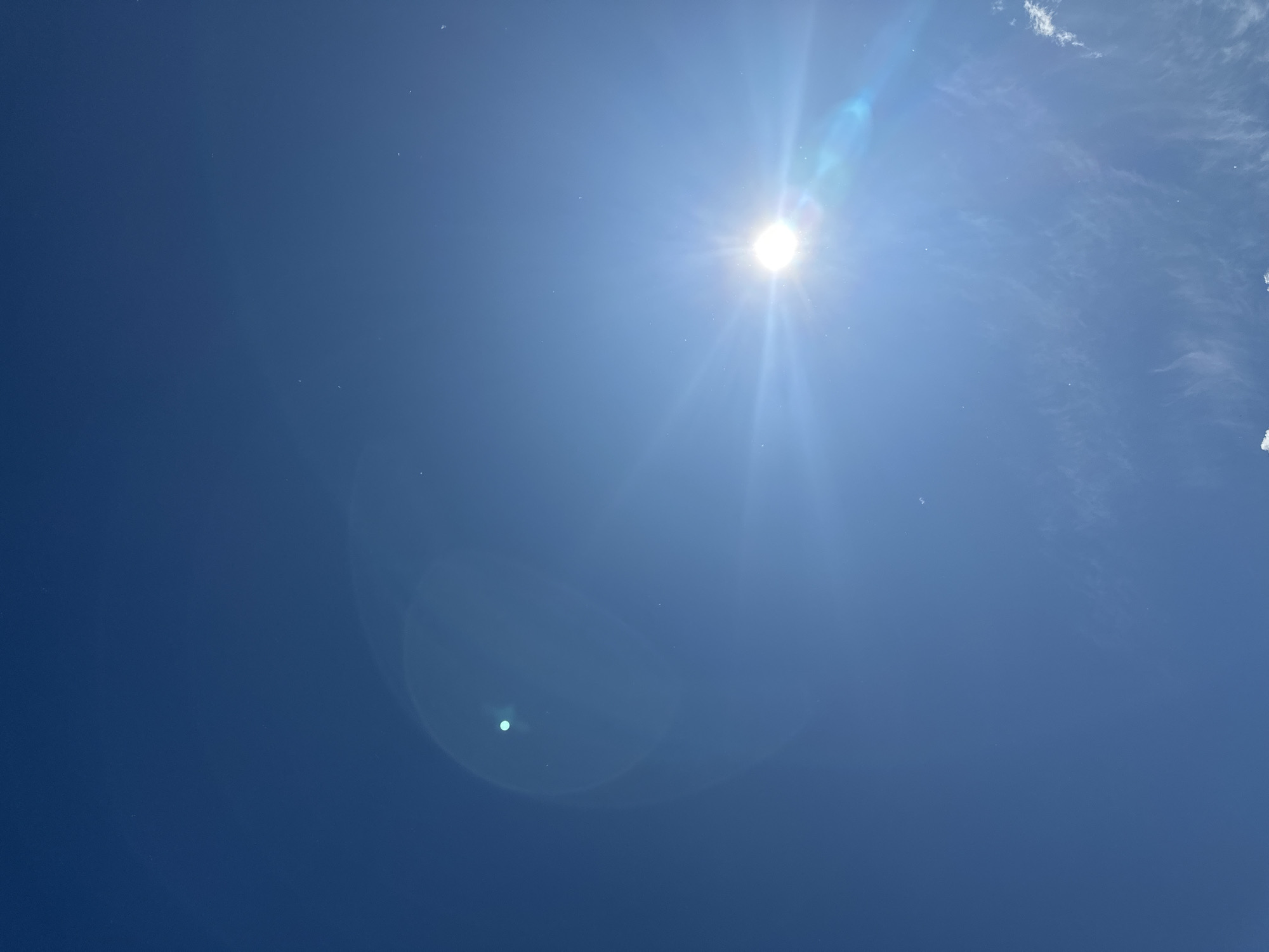 A clear, bright blue sky is depicted over Прикамский, Russia. The sun, a brilliant white orb with pronounced starburst lens flares, is positioned in the lower right portion of the frame. A distinct, circular green lens flare artifact is present in the upper left quadrant, along with other faint linear and circular lens flares traversing the sky. Numerous minute white specks or dust particles are scattered throughout the blue expanse. Minimal, wispy white cloud formations are visible in the bottom right corner. No human figures, structures, or other identifiable objects are present.