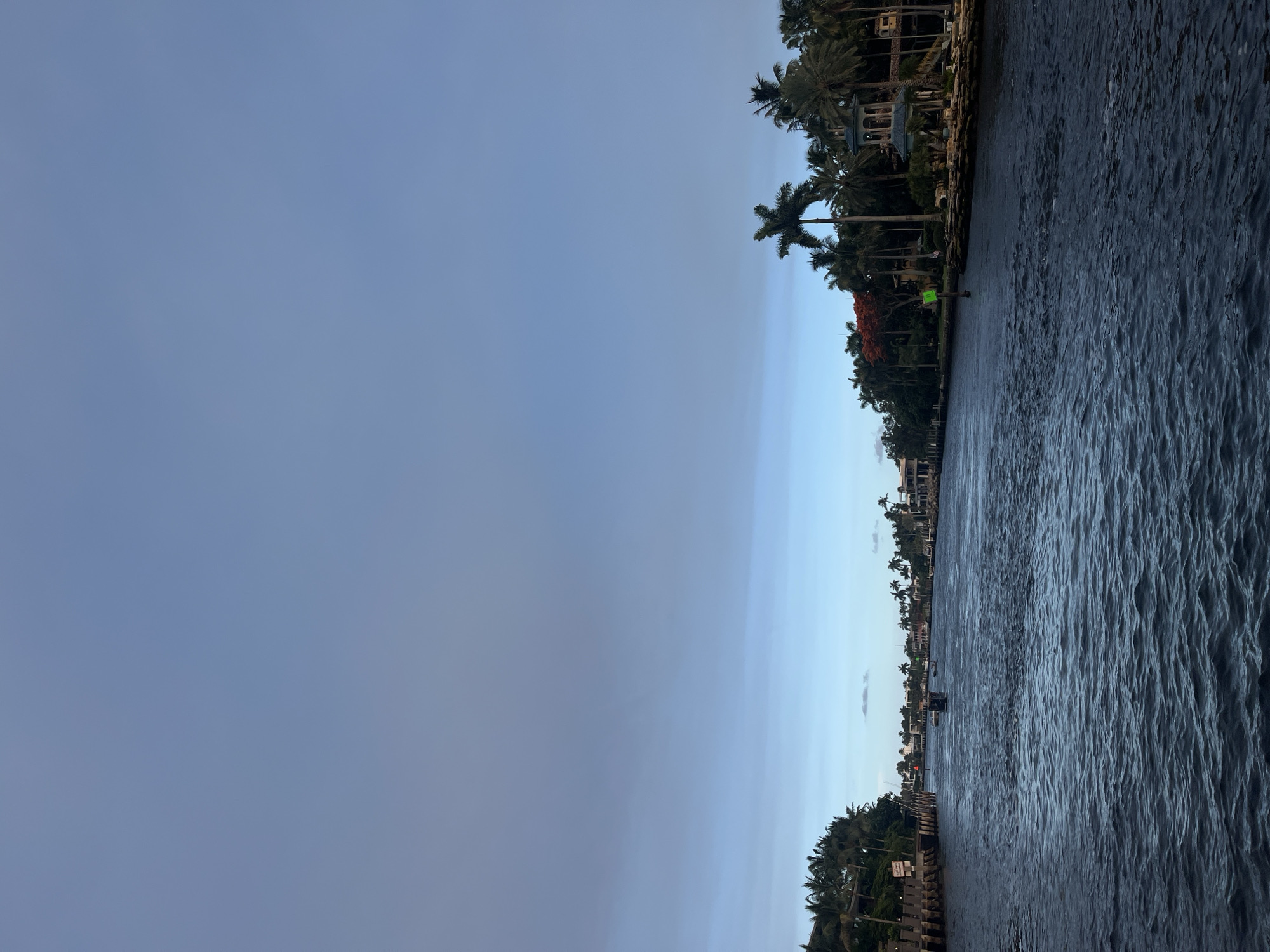 The image depicts a wide, dark blue canal in Fort Lauderdale, Florida, with the viewer positioned on the water, looking towards the horizon. The canal is lined on both sides by lush green vegetation, primarily palm trees, and luxurious waterfront homes with ornate architecture. Several boats are visible on the water, some in the distance, suggesting recreational activity. The sky above is a pale blue, transitioning into a hazy gray towards the horizon, indicating either early morning or late afternoon light. The overall scene conveys a sense of tranquility and opulence, characteristic of Fort Lauderdale's affluent canal communities.