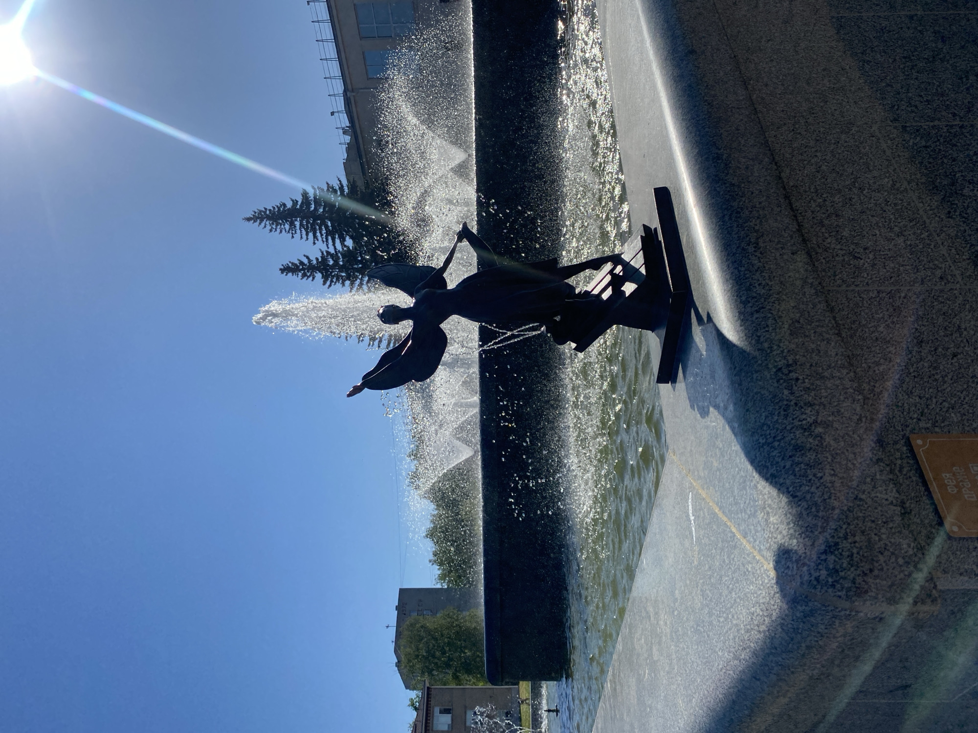 A statue of an angel standing on a bench is centrally located in a fountain in Chaikovsky, Russia. The statue and bench are silhouetted against the fountain’s water spray and the bright blue sky. The sun is in the upper left corner, creating a lens flare. Buildings and trees are visible in the background. The fountain’s edge is made of stone, with a small plaque attached to its side.