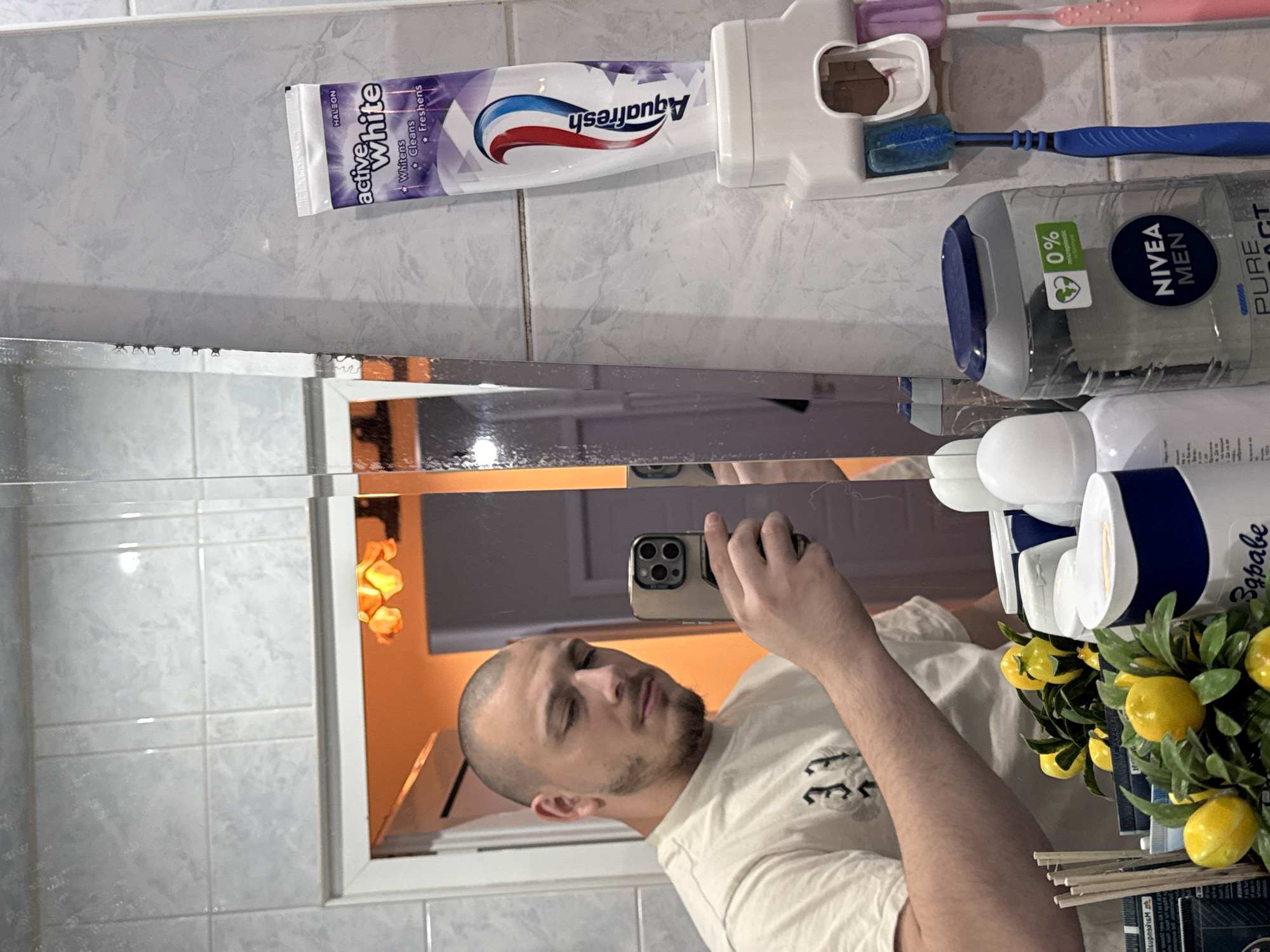 This indoor mirror selfie captures a man in a bathroom, likely in Stara Zagora, Bulgaria. The man, with a shaved head and a short beard, wears a light-colored t-shirt as he holds up an iPhone (identifiable by its multi-camera system) to take the picture. His reflection in the mirror shows a calm, neutral expression.
The bathroom features light-colored tiled walls and a mirror reflecting a warm, orange-hued light source from the room behind the man. On a shelf or counter above the mirror, various toiletries are neatly arranged. These include a tube of "Aquafresh Active White" toothpaste, with the words "Whitens," "Cleans," and "Freshens" visible, and the brand "HALEON." The toothpaste is placed in a white dispenser, next to which are a blue and a pink/purple toothbrush. A bottle of "NIVEA MEN PURE IMPACT" shower gel or shampoo is also present, marked with "0% microplastic." Another white bottle has the partial text "Здраве" (Bulgarian for "Health"). A small decorative plant with artificial yellow lemons and some diffuser sticks add a touch of decor to the setting.