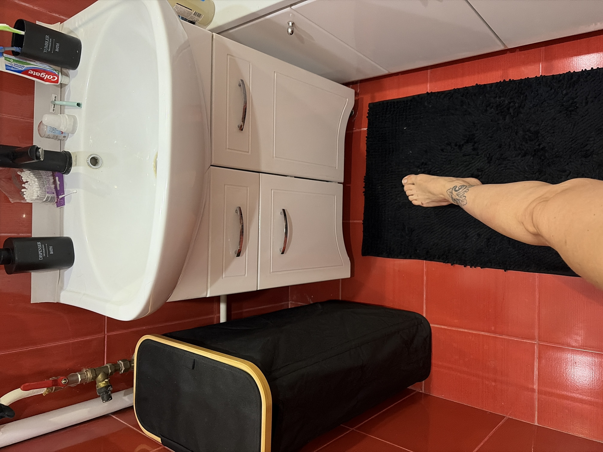 This is a medium-shot, high-angle, indoor photograph of a bathroom. The bathroom has bright red tiled walls and floor. A white porcelain sink is mounted on a white cabinet with silver handles. On the sink's edge, there is a black soap dispenser, cotton swabs, a razor, a tube of Colgate toothpaste, and a black cup holding toothbrushes. A black laundry hamper with a wooden rim is positioned to the left of the sink. In the foreground, a person's bare left leg and foot are visible, standing on a black bath mat. The foot has a tattoo on the ankle. The scene suggests a domestic setting in a bathroom.