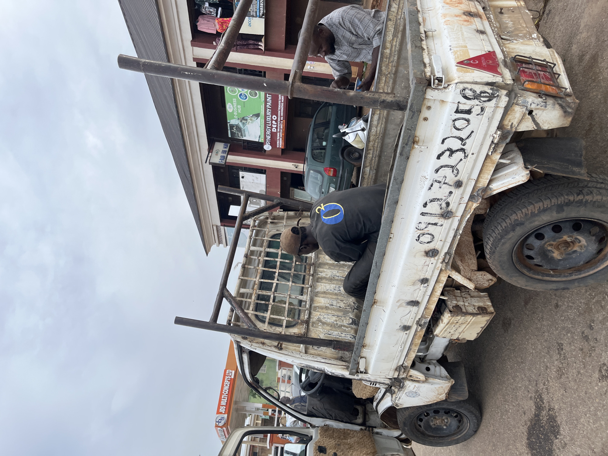 This media file shows a white pickup truck with a cage-like structure on its bed, parked on a street in Gura Top, Nigeria. The truck appears weathered and has some graffiti on its side, including a phone number and the text "0912.7232058". Two individuals are visible in and around the truck. One person, wearing a plaid shirt, is seen from the chest up, leaning into the truck bed and appearing to be working on something. Another person, wearing a black t-shirt with a blue and yellow circular logo, is seated in the truck bed.
In the background, a building with shops is visible. One shop has a sign advertising "SYNERGY LUXURY PAINT" and "E-BUSINESS HUB". Another sign reads "JDS MULTI-CONCEPTS LTD". A green car and a motorcycle are parked in front of the shops. The sky is overcast, suggesting a cloudy or possibly rainy day. The overall scene depicts a commercial street with a focus on the truck and the activity surrounding it.