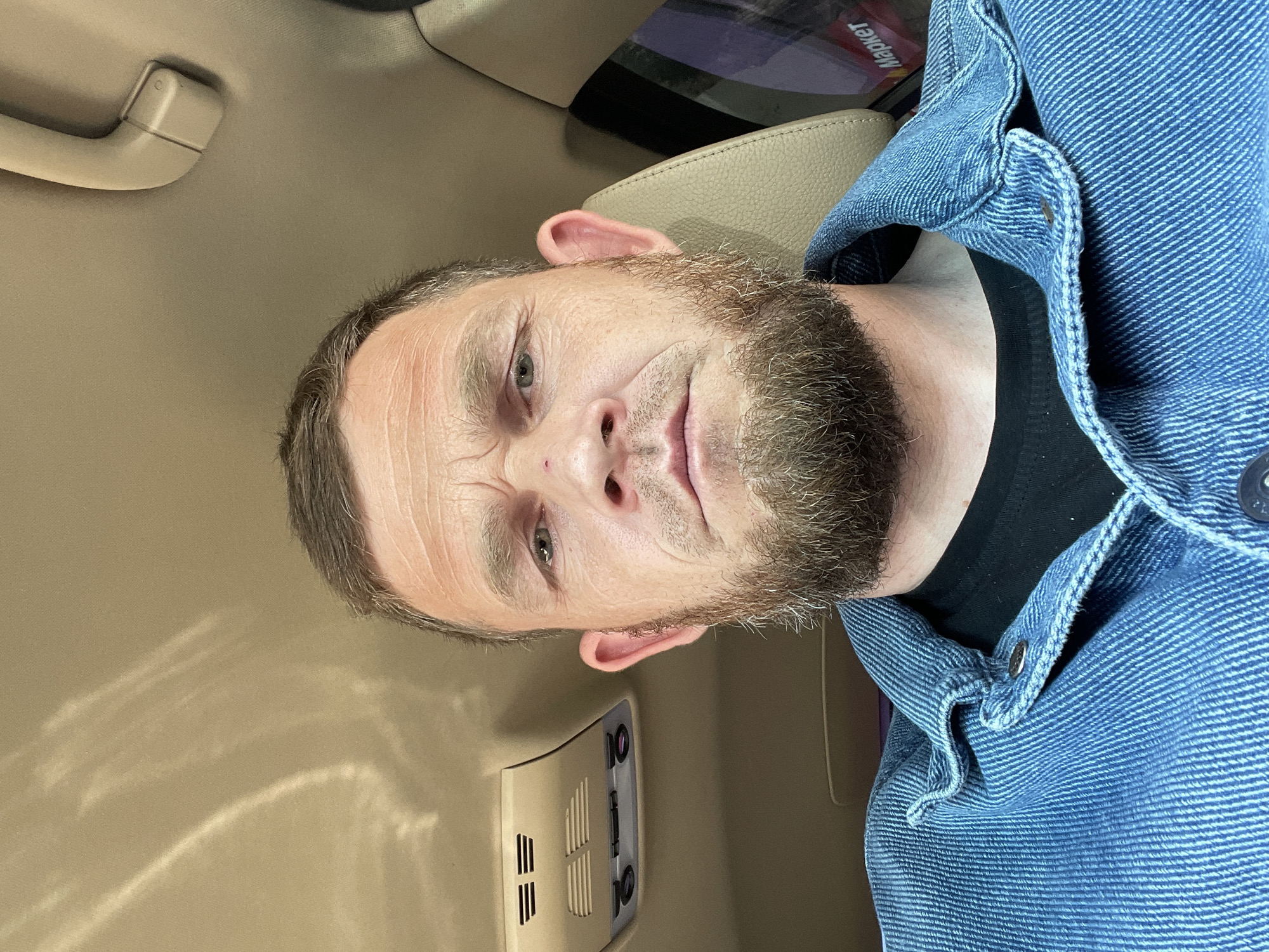 A close-up, slightly angled portrait shows a middle-aged Caucasian man with a beard, possibly in his late 30s to early 50s, inside what appears to be a vehicle. The man has light skin, graying hair, and a beard of mixed light and dark hairs. He is wearing a blue denim jacket over a black shirt. The interior of the vehicle is beige or light tan, visible on the ceiling and parts of the dashboard. A handle is visible near the top left, and a control panel with vents and buttons is seen near the bottom left. A reflective object with the word "Маркет" is partially visible at the top, suggesting the location might be in Russia, possibly near a "Market" (translated). Given the request for the city, and knowing the Cyrillic text, Krasnodar is a reasonable conjecture, but this is based on limited visual cues. No specific interaction or action is taking place in the frame.