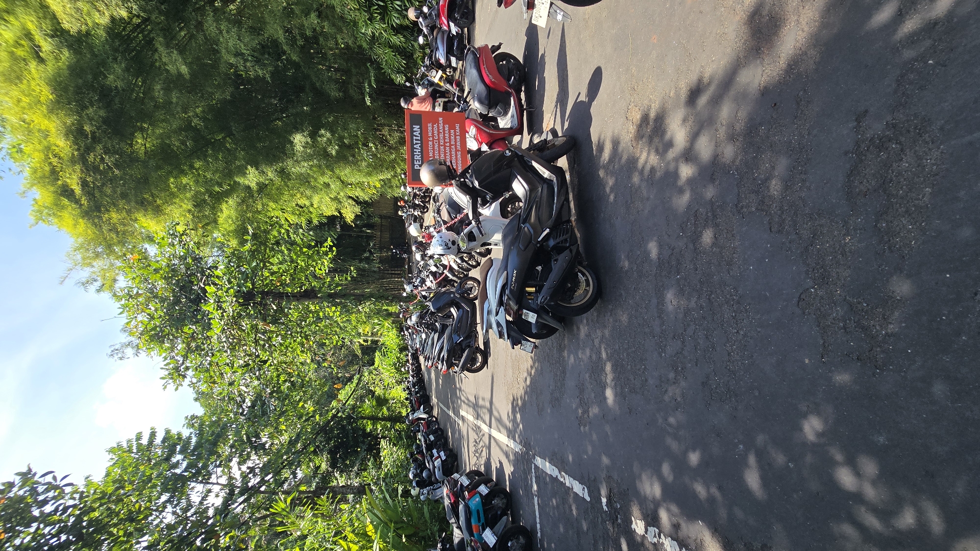 The image shows a parking lot filled with motorcycles and scooters in Celuk, Indonesia. The lot is paved with asphalt and marked with white lines to delineate parking spaces. The motorcycles are parked closely together, with varying colors and models visible. A sign in Indonesian warns motorists about securing their vehicles against theft, stating that the parking management is not responsible for losses. Lush green trees and foliage surround the parking lot.