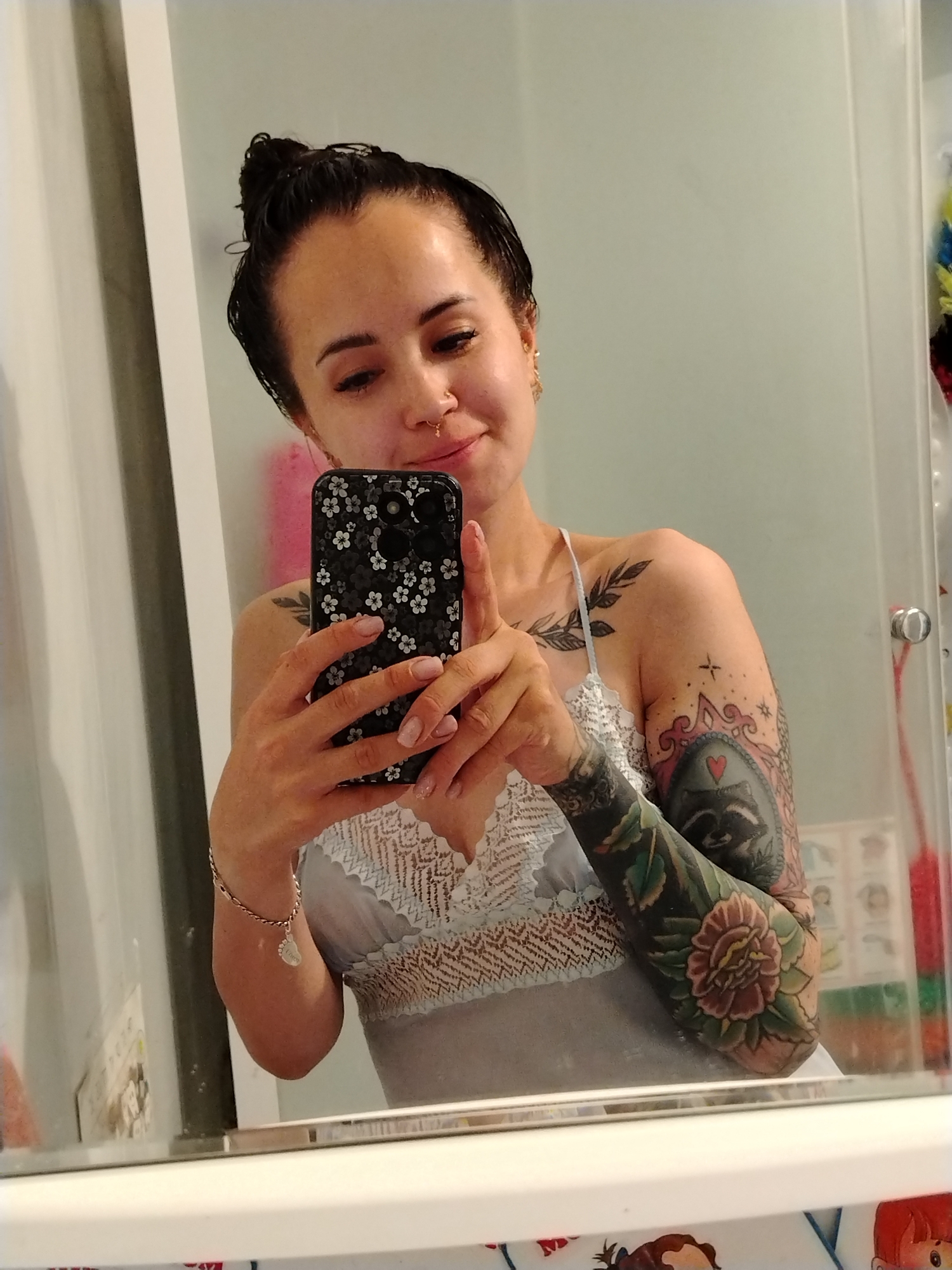 A woman with dark hair pulled into a bun is taking a selfie in a bathroom mirror. She is wearing a light blue lacy camisole. She has multiple tattoos on her arms and shoulder, including a raccoon, a flower, and some leafy branches. She is also wearing a bracelet on her left wrist with a small foot-shaped charm. Her phone has a black floral case. The background is a plain wall, with a shower curtain and some shelving visible on the right side. The photo was taken in Noviy, Russia.