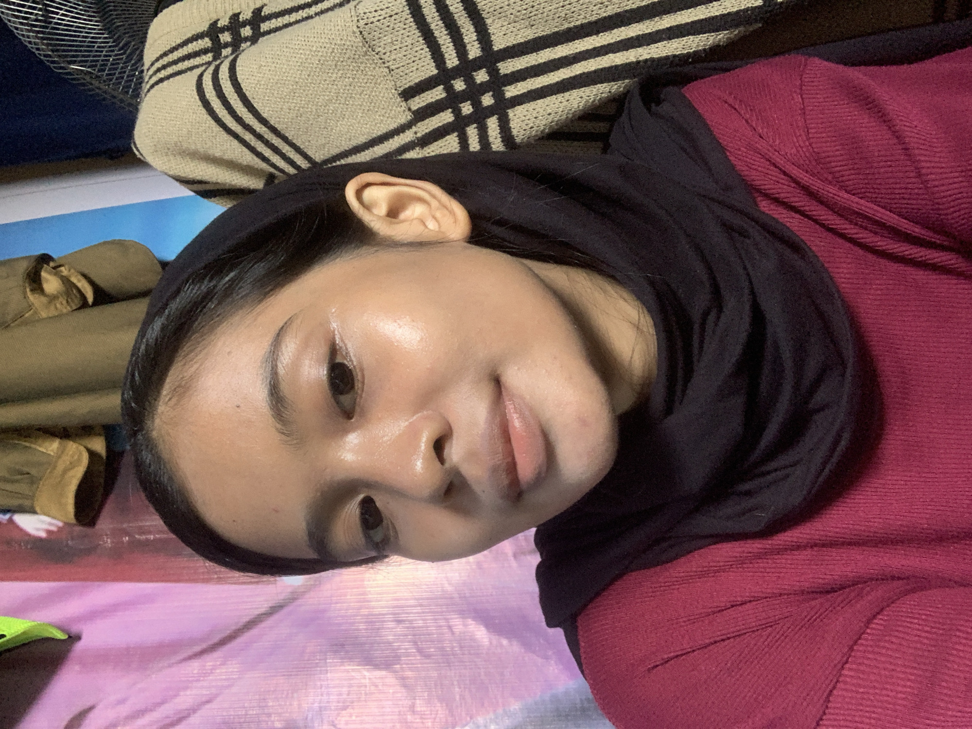 A young woman wearing a black hijab and a maroon shirt is pictured indoors. The background consists of a pink textured fabric, a blue wall with what appears to be clothing hanging on it, and a patterned blanket. The woman has dark hair and is looking towards the camera with a subtle smile. Her skin has a dewy appearance, possibly due to makeup or lighting. The image is a close-up shot, focusing primarily on her face and upper body. The lighting suggests an indoor setting with a soft, warm glow. There is no indication of the specific location in Sungai Meriam, Indonesia, within the image itself.