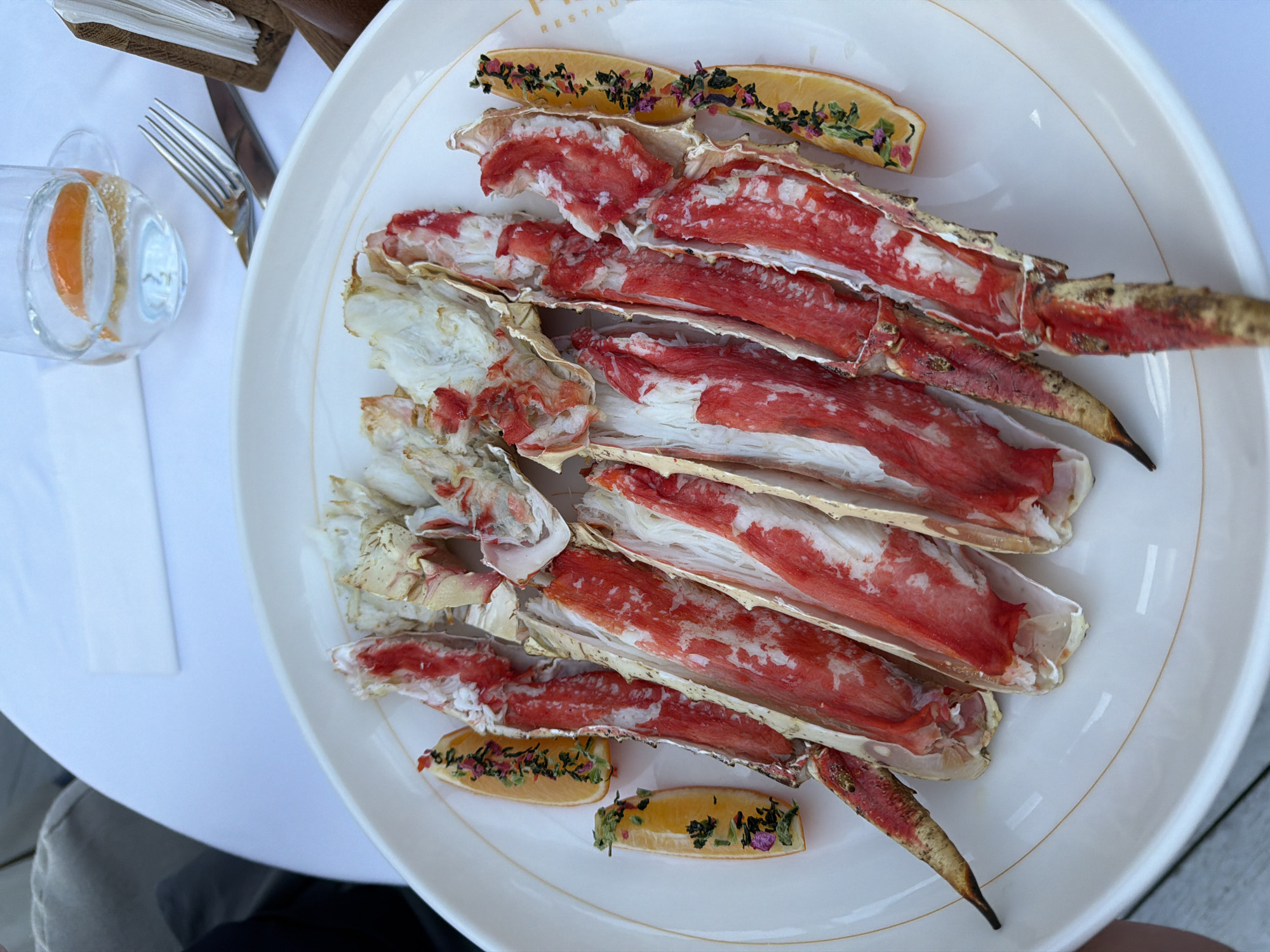 The primary subject of this image is a large, white oval plate generously filled with cooked crab legs, likely King Crab or Snow Crab, artfully arranged in a dining setting. The crab legs are split lengthwise, revealing the bright red and white tender meat within their shells. Several lemon or lime wedges, garnished with a mixture of dried herbs and small, colorful flower petals, are placed on the plate as an elegant accompaniment.
The plate rests on a pristine white tablecloth, suggesting a formal or upscale dining experience. To the left, a clear glass contains an orange slice, and a silver fork and knife are visible beside it. A wooden napkin holder is partially seen in the top left corner. The scene is illuminated by bright, natural light, indicating a daytime meal. Part of a person's leg or arm is visible at the bottom left, suggesting someone is seated at the table enjoying the meal. The word "RESTAURA" is partially visible on the rim of the plate, confirming a restaurant setting. This culinary moment takes place in Moscow, Russia. The overall impression is one of a luxurious and appealing seafood meal served with careful presentation.