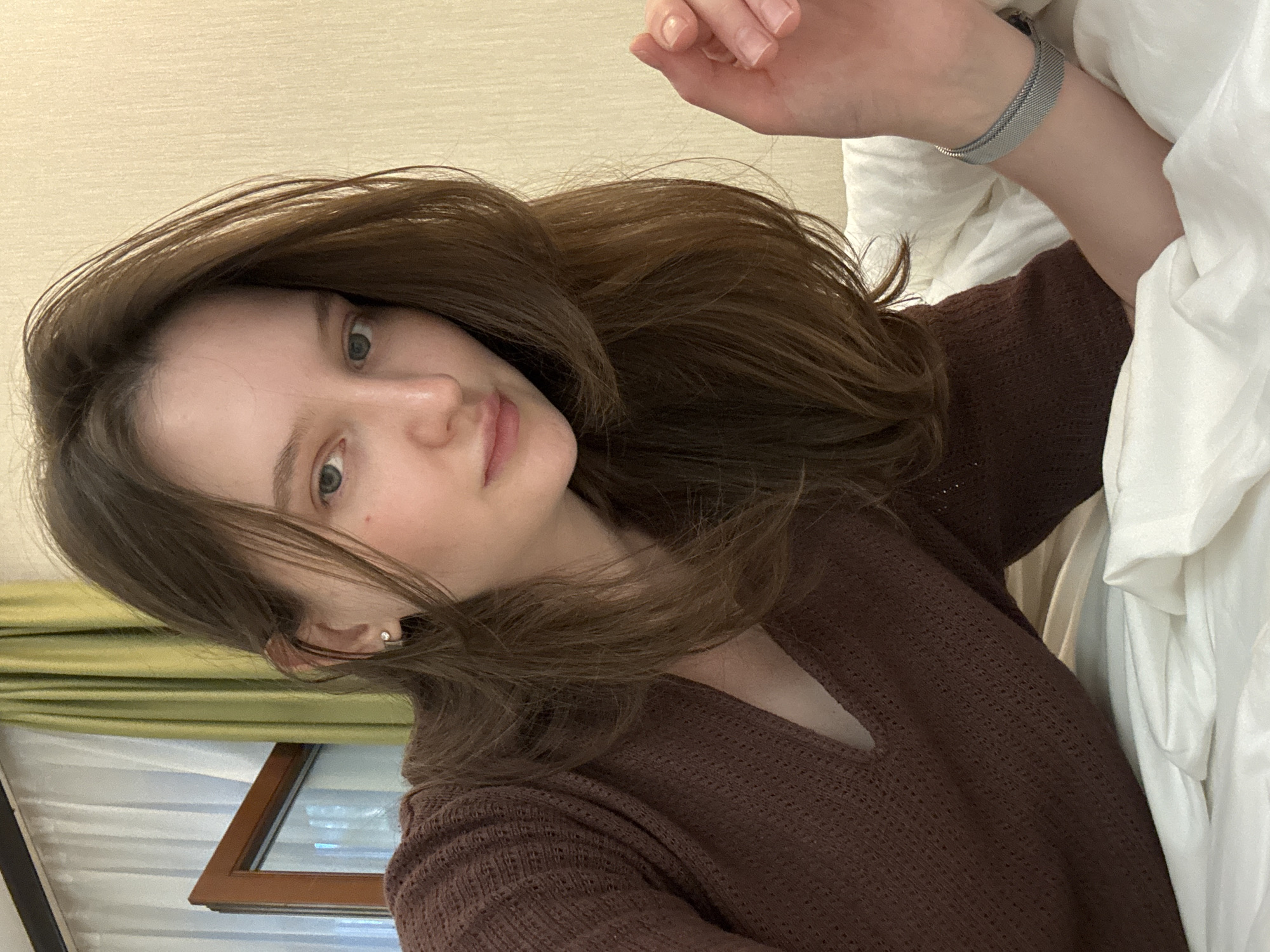 This media file features a young woman lying in bed. She has long, brown hair that is casually styled and falls around her face and shoulders. She is wearing a brown, textured sweater. Her eyes are open, and she appears to be looking towards the camera. A silver mesh watch is visible on her left wrist.
The setting appears to be indoors, likely a bedroom, with white bedding prominently featured. A window with green curtains and a view of what looks like a window frame with a reflection is visible in the background. The lighting suggests it could be daytime, possibly morning or afternoon. There is no visible text in the image. The overall mood is relaxed and intimate, as if capturing a candid moment.