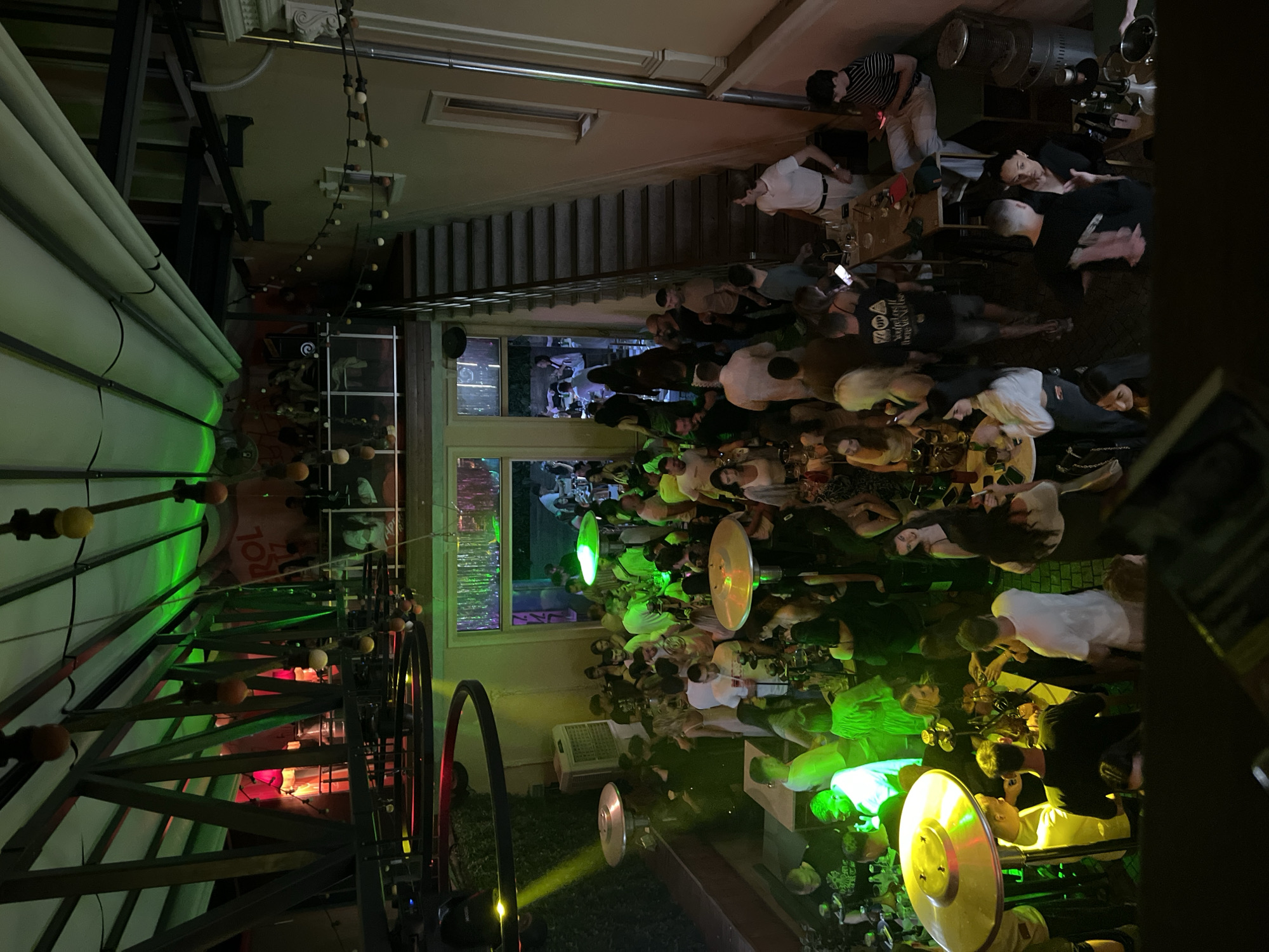 The image shows a crowded outdoor patio in an urban area, possibly a bar or restaurant. The location is unspecified in the city of Unknown, Unknown. People are densely packed at tables, some standing, suggesting social interaction and likely eating or drinking. The scene is illuminated by both overhead lights and possible patio heaters, some glowing yellow. There are visible architectural features, including stairs and a window frame. A structure possibly related to an awning or decorative element dominates the left side of the frame, featuring a red and green colour scheme. The environment has a lively, bustling atmosphere, likely during evening hours.