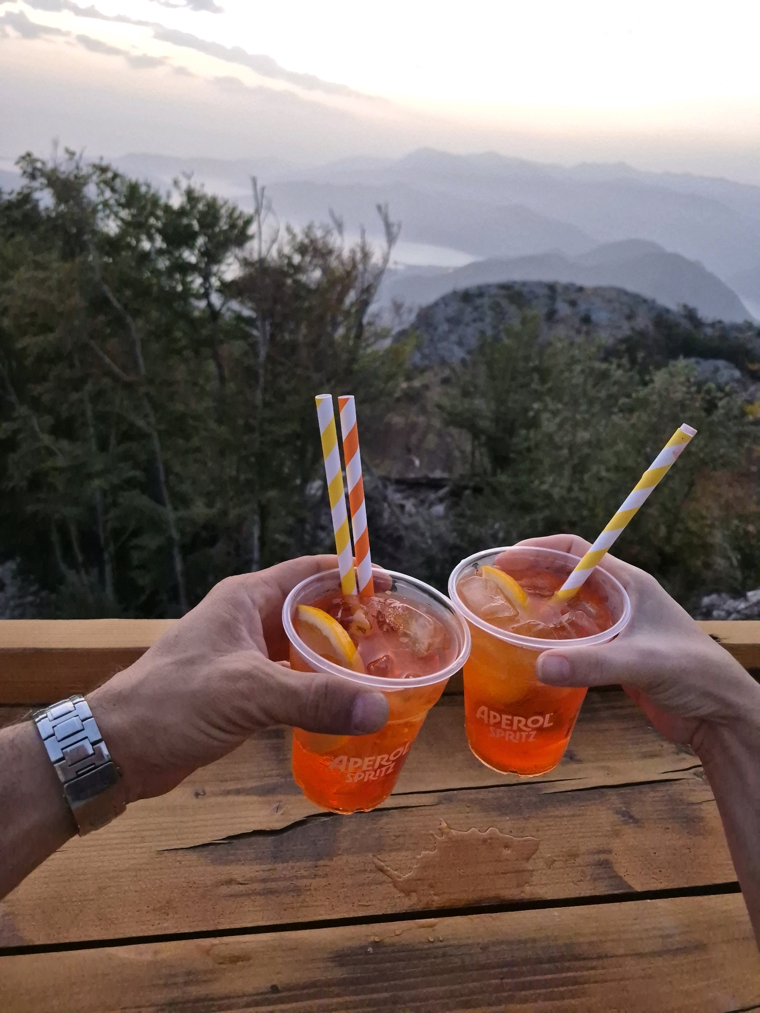 Two clear plastic cups filled with an orange beverage and ice are held up. Both cups have a slice of orange on the rim and a striped paper straw. The cups are branded with "APEROL SPRITZ". The cups are held by two individuals' hands. One hand on the left wears a silver watch. The cups are positioned in the foreground, resting on a wooden table. In the background, there are green trees and a hazy mountainous landscape with a body of water visible in the distance. The sky above is a light pink and orange hue, suggesting it is dusk or dawn. The location is identified as a viewpoint in Kuk, Montenegro.