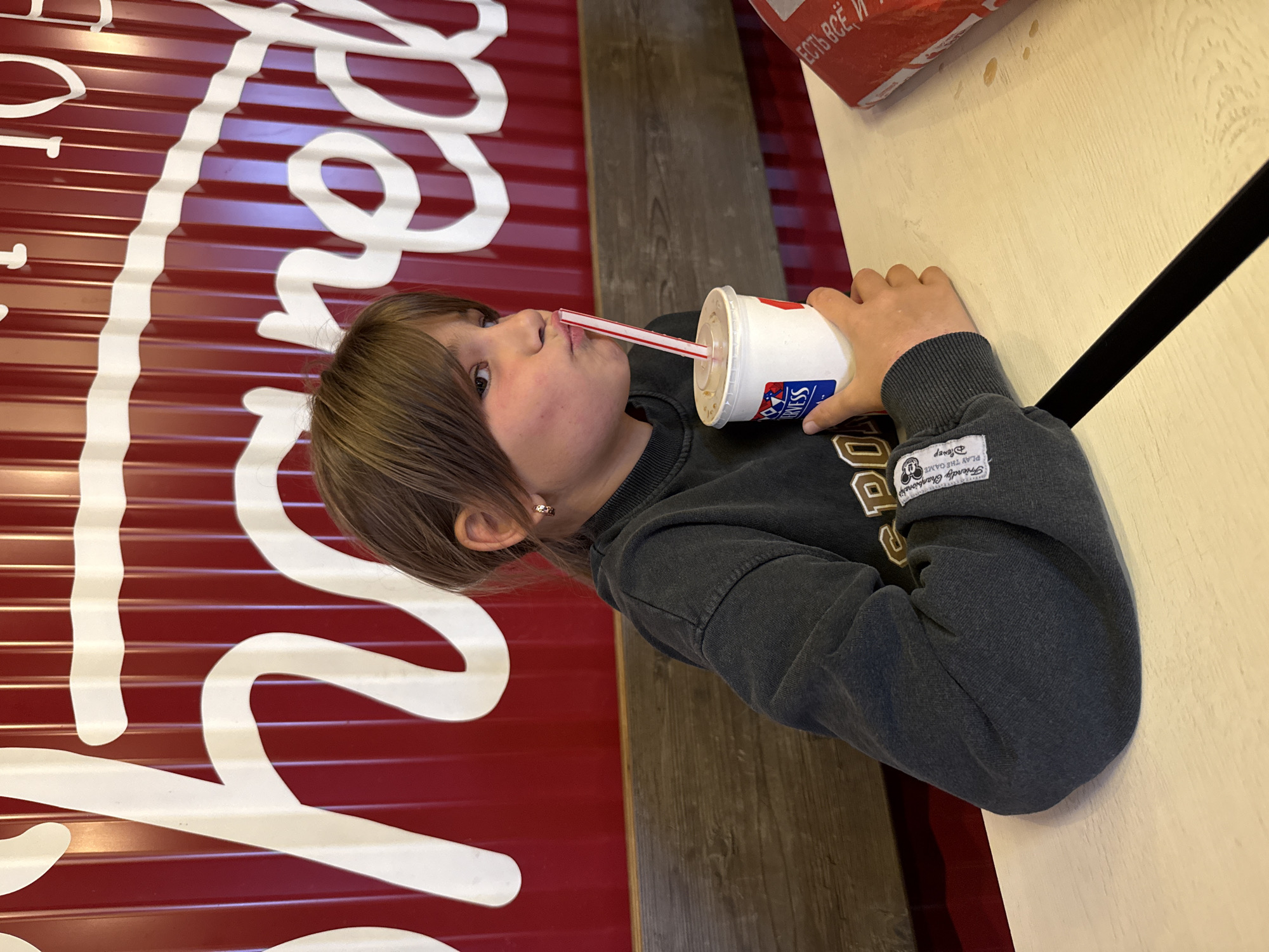 In the city of Ignatovo, Russia, a young person with brown hair is seated at a table, holding a white cup with a red and white striped straw and drinking from it. The person is wearing a dark gray sweatshirt. The background features a red corrugated wall with white wavy lettering. The table appears to be made of light-colored wood.