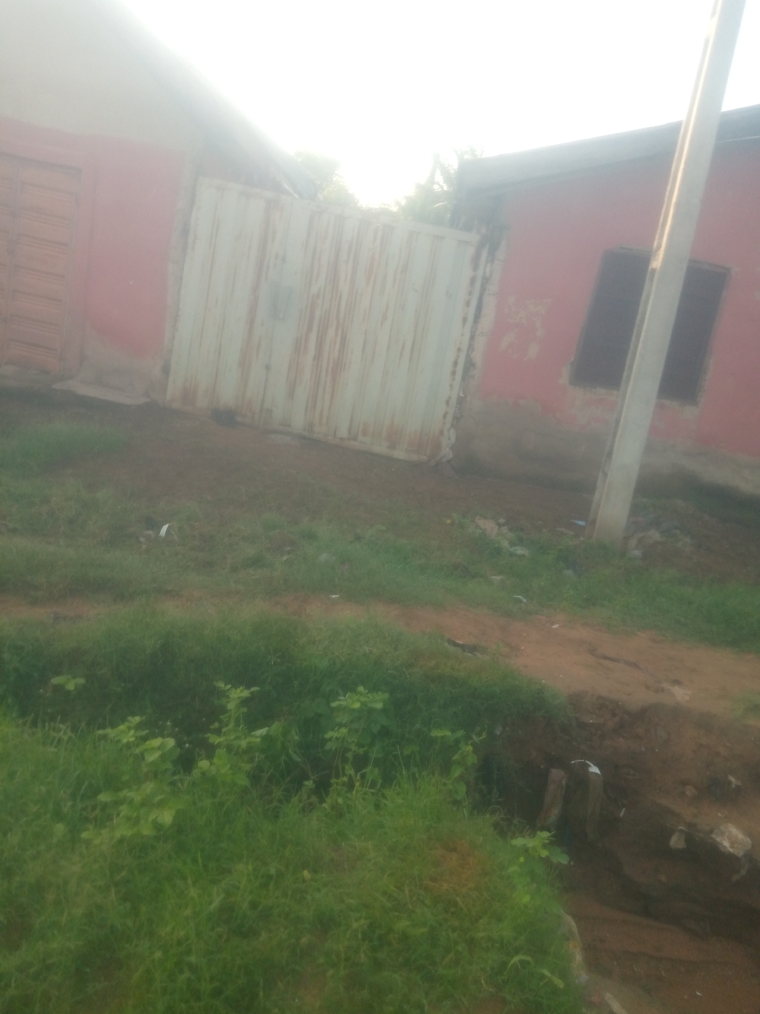 The image depicts a street scene in Jauro Yinu, Nigeria, featuring a red building with a large, corrugated metal gate. To the left of the gate, a section of the building shows a brown door. The building has at least one dark-colored window visible on the right side. A prominent, light-colored utility pole stands near the right edge of the frame, extending upwards. The ground in front of the building is a mixture of dirt and patchy grass, with some visible debris. The lighting suggests it might be daytime, though the sky appears bright and possibly hazy or overexposed. There are no people or specific actions visible in the image.