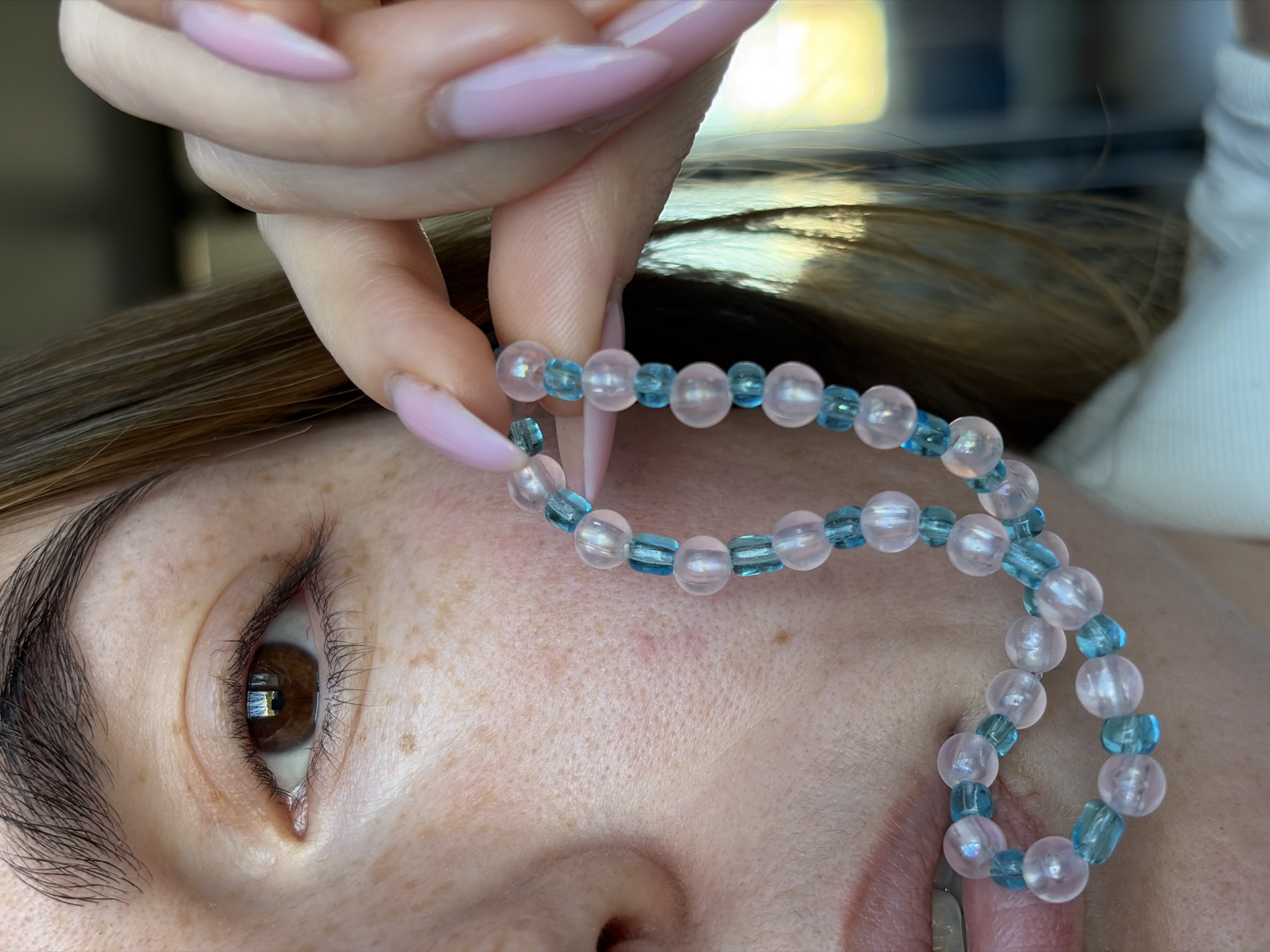 A close-up shot depicts a person lying down, with a hand holding a pink and blue beaded bracelet over their face. The person's eye is visible, as are freckles on their skin. The fingernails of the hand holding the bracelet are painted light pink. The background is blurry and indistinct, but there's some light and the vague suggestion of a white surface. The scene doesn't explicitly contain information that would allow one to determine a specific location, such as Izhevsk, Russia.