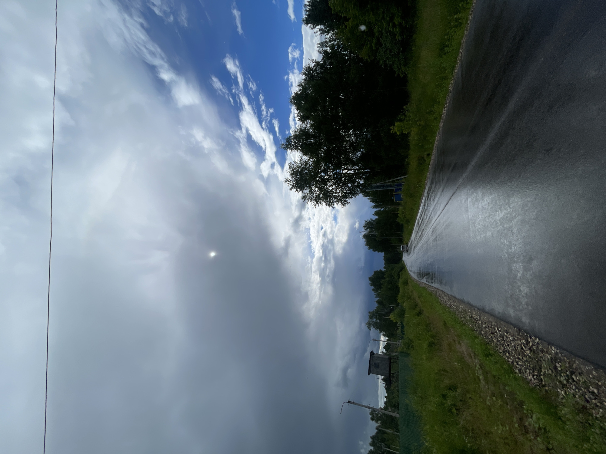 The image depicts a wet asphalt road curving to the right, reflecting the sky. The road is bordered by a gravel shoulder and lush green grass. On the left side of the road, a tall wooden structure resembling a hunting blind or observation tower is visible, accompanied by a utility pole. A green metal fence runs along the edge of the property. The sky is dramatic, with a mix of dark, heavy storm clouds on the left and bright blue sky with scattered white clouds on the right. The sun is partially obscured by the clouds, creating a bright, diffuse halo. Dense green trees line the right side of the road, forming a dark treeline. A single white vehicle is visible in the distance on the road. The overall impression is of a rural or semi-rural setting near Zarechny, Russia, experiencing inclement weather.