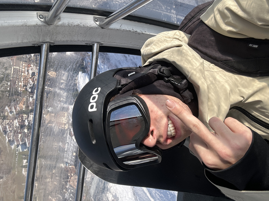 A young adult male, presumed to be a snowboarder, is situated inside a gondola lift cabin, ascending over a snow-covered landscape in La Clusaz, France. The individual wears a black "POC" brand ski helmet, black reflective ski goggles that obscure his eyes, a black balaclava covering his lower face and ears, and a light beige winter jacket with black shoulder accents, one of which displays a "NIKE SB" logo. He is smiling, showing his upper teeth, and holding his left index finger to his left cheek, pointing towards his mouth. A black snowboard is partially visible positioned vertically behind his left shoulder. The interior of the gondola cabin shows metal framing and clear, scratched window panels. Outside the window, a snowy mountain slope is visible immediately below, transitioning into a village composed of numerous chalet-style buildings nestled among snow-dusted trees in the mid-distance. The scene is brightly illuminated by direct sunlight.