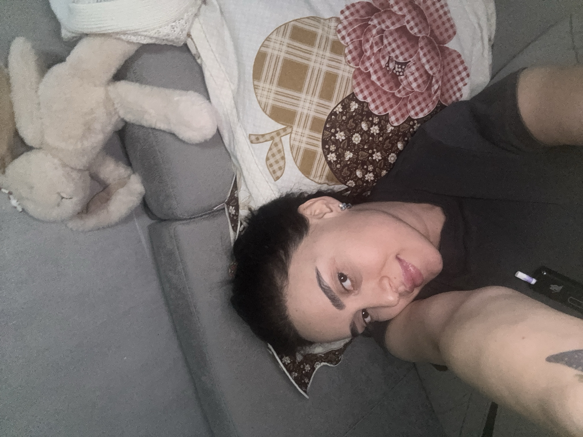 A woman lies on a gray sofa next to a white stuffed rabbit and a pillow with an apple and flower pattern. The woman has dark hair, dark eyebrows, and fair skin. She is wearing a dark t-shirt. Her head rests on the floral-trimmed pillow, and her arm is extended. A black object with a white cylindrical component is near her arm. The environment appears to be an interior setting. The location cannot be determined from the image.