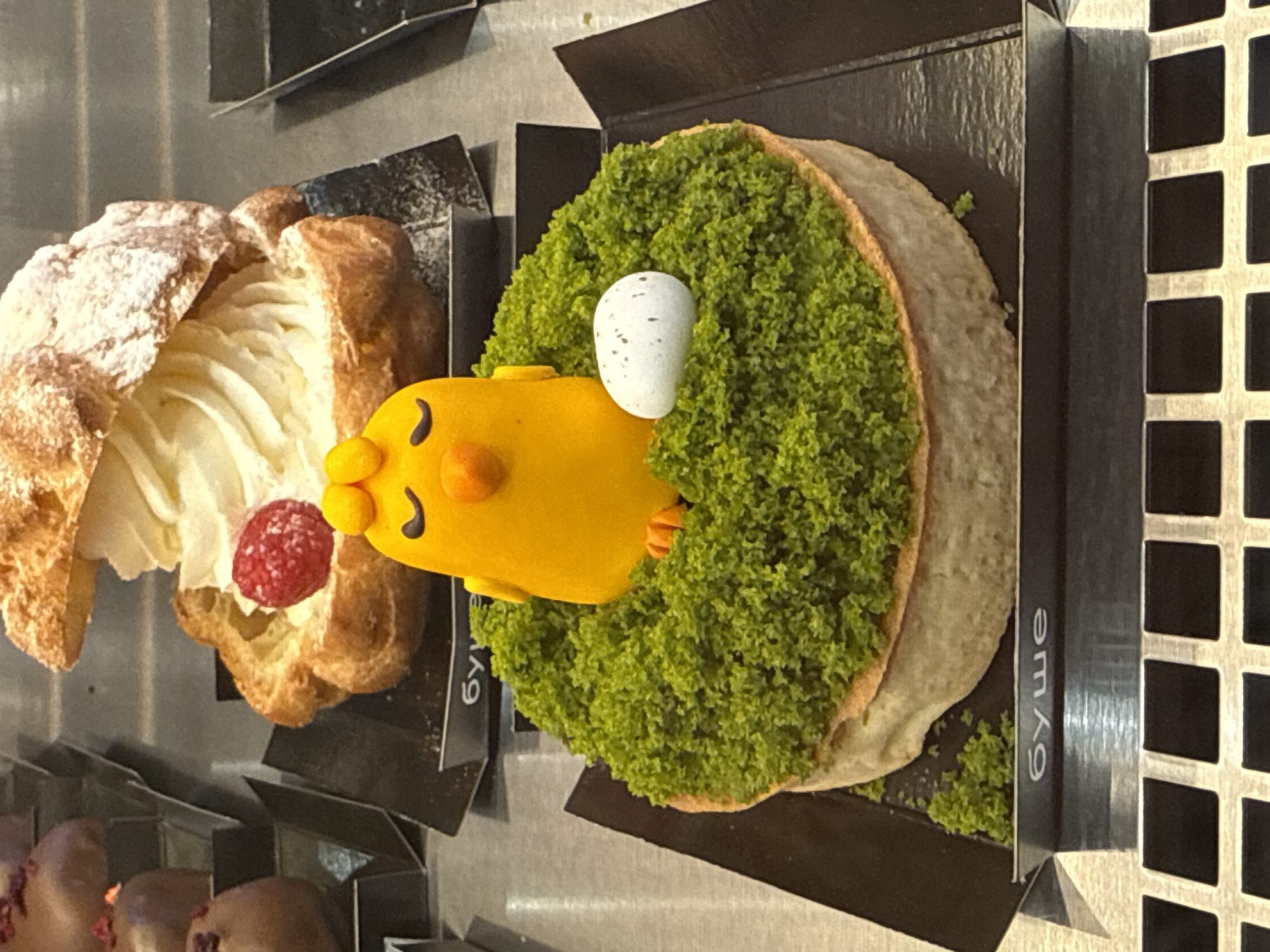 The image shows two pastries, one a cream puff with powdered sugar and a raspberry, and the other a chick-shaped cake with green topping and an egg detail. Both pastries are displayed in individual black containers with the name "Буше" printed on the sides, suggesting they are from a bakery or cafe chain called Bushe. The setting is likely a pastry display case, possibly in a Bushe location in Saint Petersburg, Russia, where the chain is known to exist. Other pastries are visible in the display below. The lighting seems to be from above, as there is some shine reflecting off the surfaces.