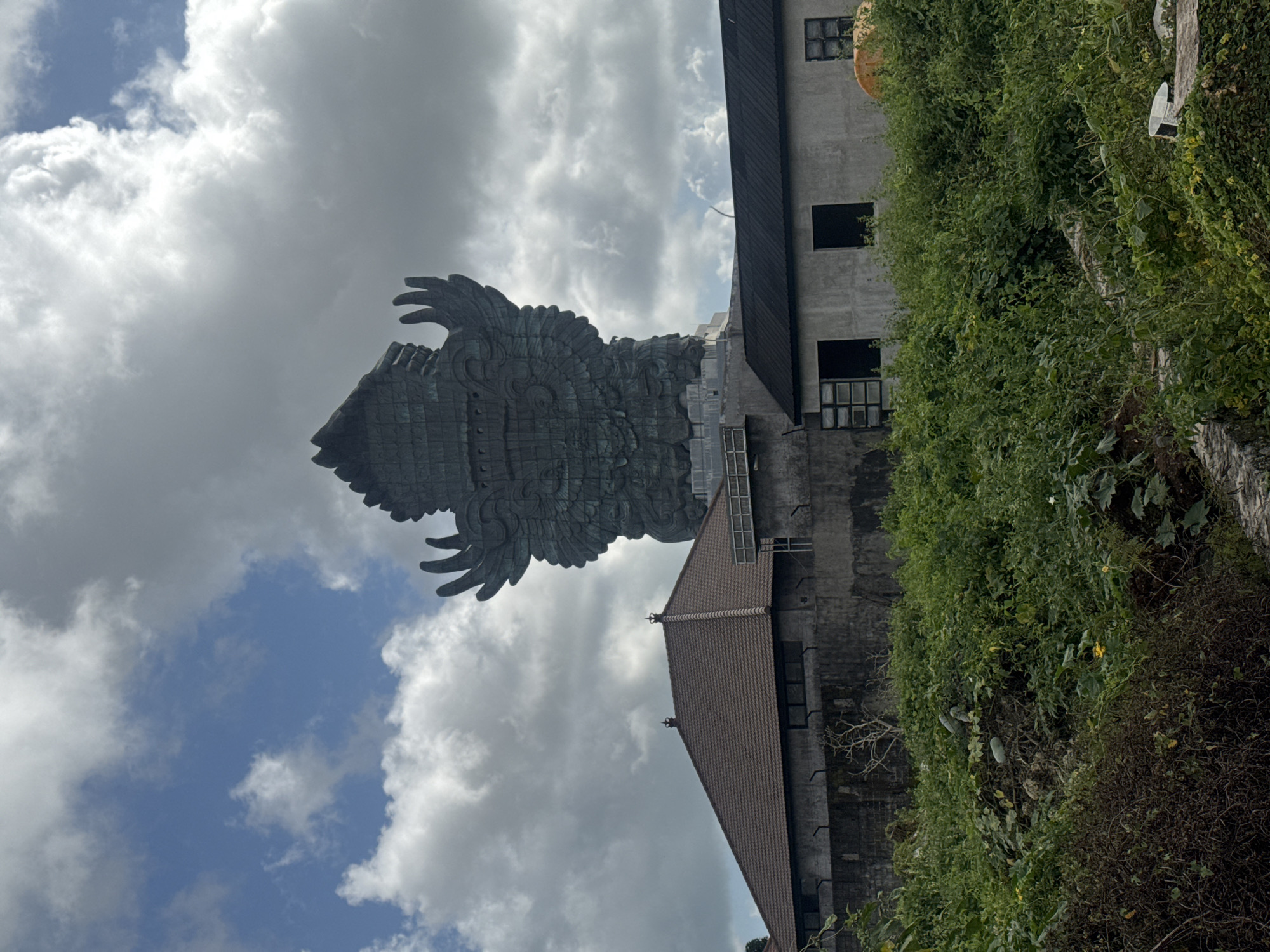 In Kuta Selatan, Indonesia, a massive, dark-colored statue of Garuda, a mythical bird-like creature with outstretched wings, dominates the background against a sky with scattered white clouds. The statue, appearing to be made of metal or stone, is incredibly detailed and imposing. In the middle ground, a building with a brown tiled roof and dark windows is visible, constructed with a grey concrete wall. To the right of this building, another, simpler structure with a dark roof and grey concrete walls and windows is present. The foreground is filled with lush green vegetation, including leafy plants and some vines, with a few pale green gourds visible amidst the foliage. The overall scene is dominated by the colossal statue and the natural and man-made structures in front of it.