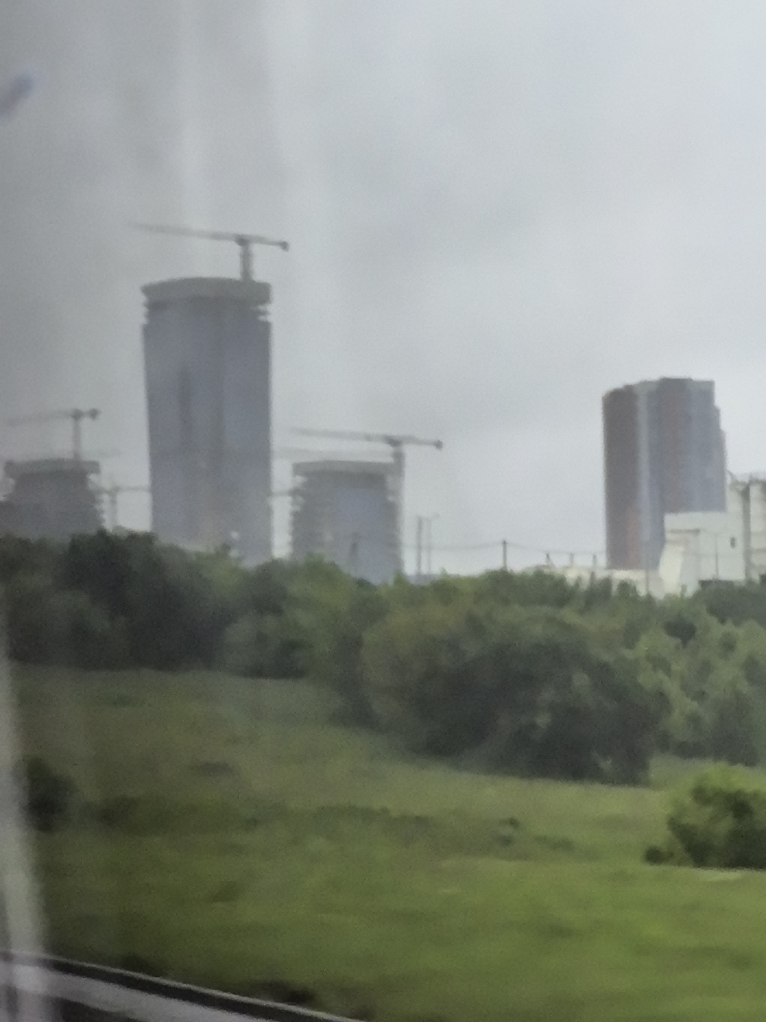 A photograph taken from a moving vehicle shows a city skyline under a cloudy sky. The foreground features a grassy hill dotted with trees. In the background, several tall buildings are under construction, identifiable by the cranes and scaffolding visible at their tops. One prominent, completed skyscraper stands to the left of center, appearing as a slender, glass-fronted structure. To the right, another completed building features a rounded facade. The overall atmosphere is somewhat muted due to the overcast weather.