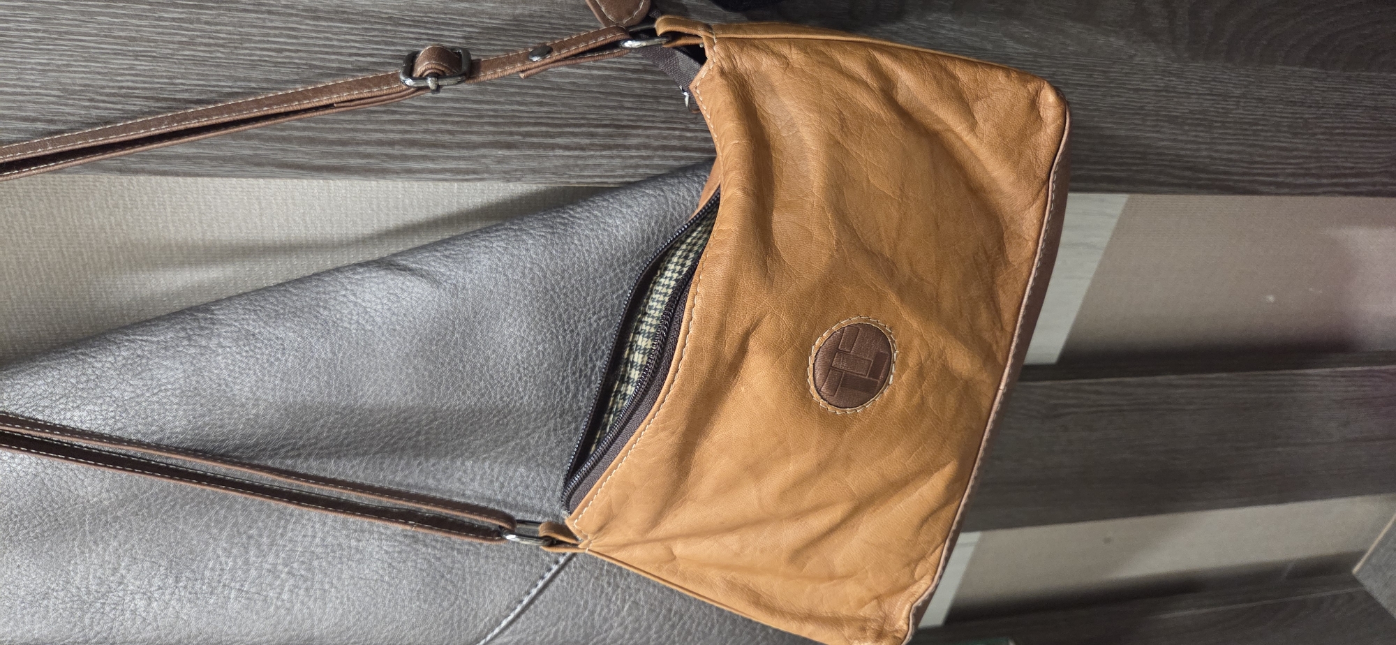 A brown leather handbag with a dark brown zipper and brown leather strap is displayed against a textured grey surface, possibly a wall or piece of furniture. The handbag has a circular logo with a dark brown emblem on its front. The background appears to be a wooden surface with vertical lines. The image is a close-up of the handbag, with no people or other significant objects visible. The lighting suggests an indoor setting. There is no information or visual evidence to suggest this image is located in the city of Lipetsk, Russia.