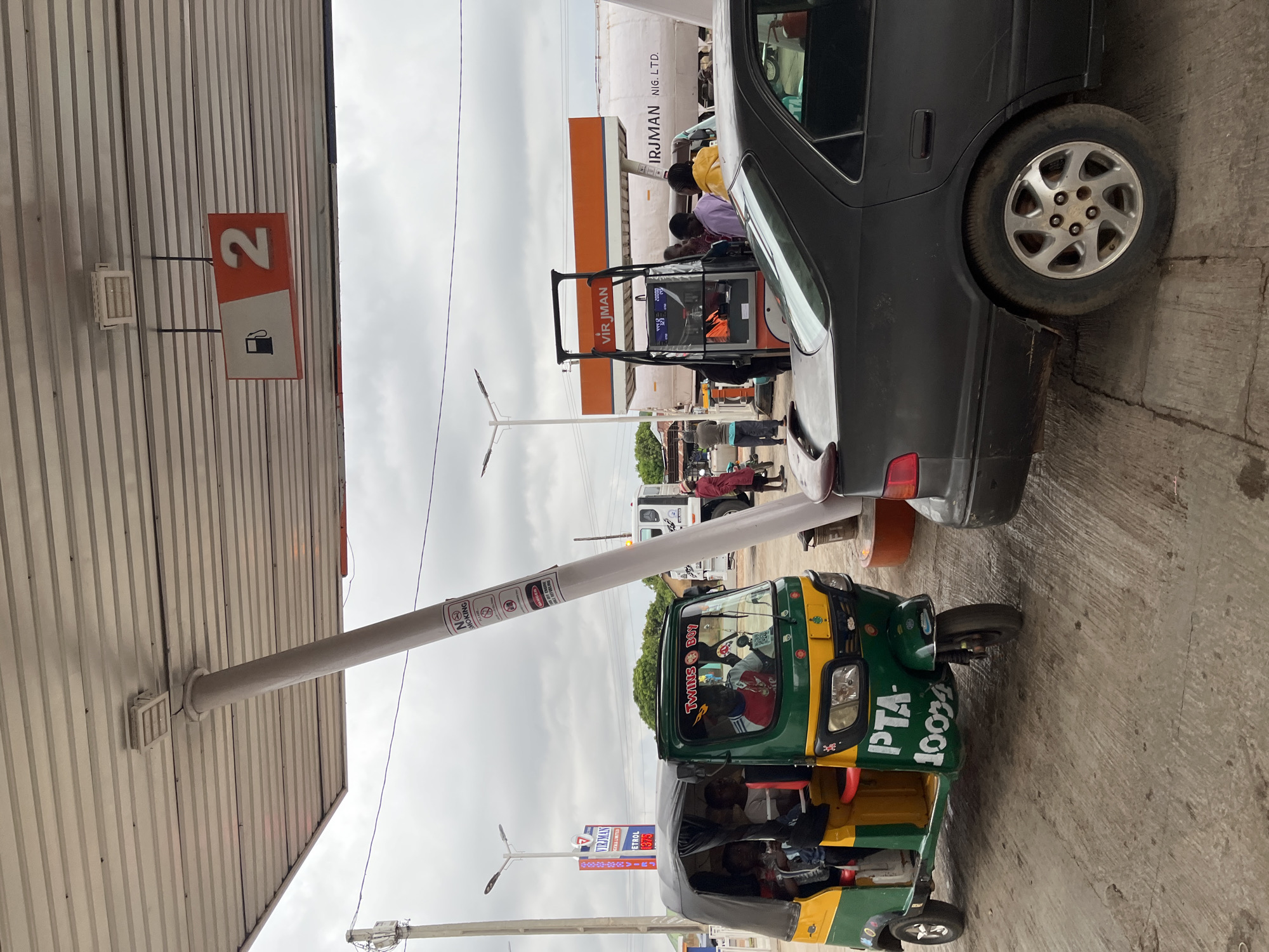 The image depicts a scene at a Virjman petrol station in Jos, Nigeria. A green and yellow "keke," or auto-rickshaw, with the registration "PTA 10004," is parked under the station canopy. Next to it is the rear of a black car. People are filling their cars at the Virjman pumps, and the price of petrol is displayed as 1375. A sign indicates "2" for the petrol pump and a "no smoking" sign is visible on a pole.