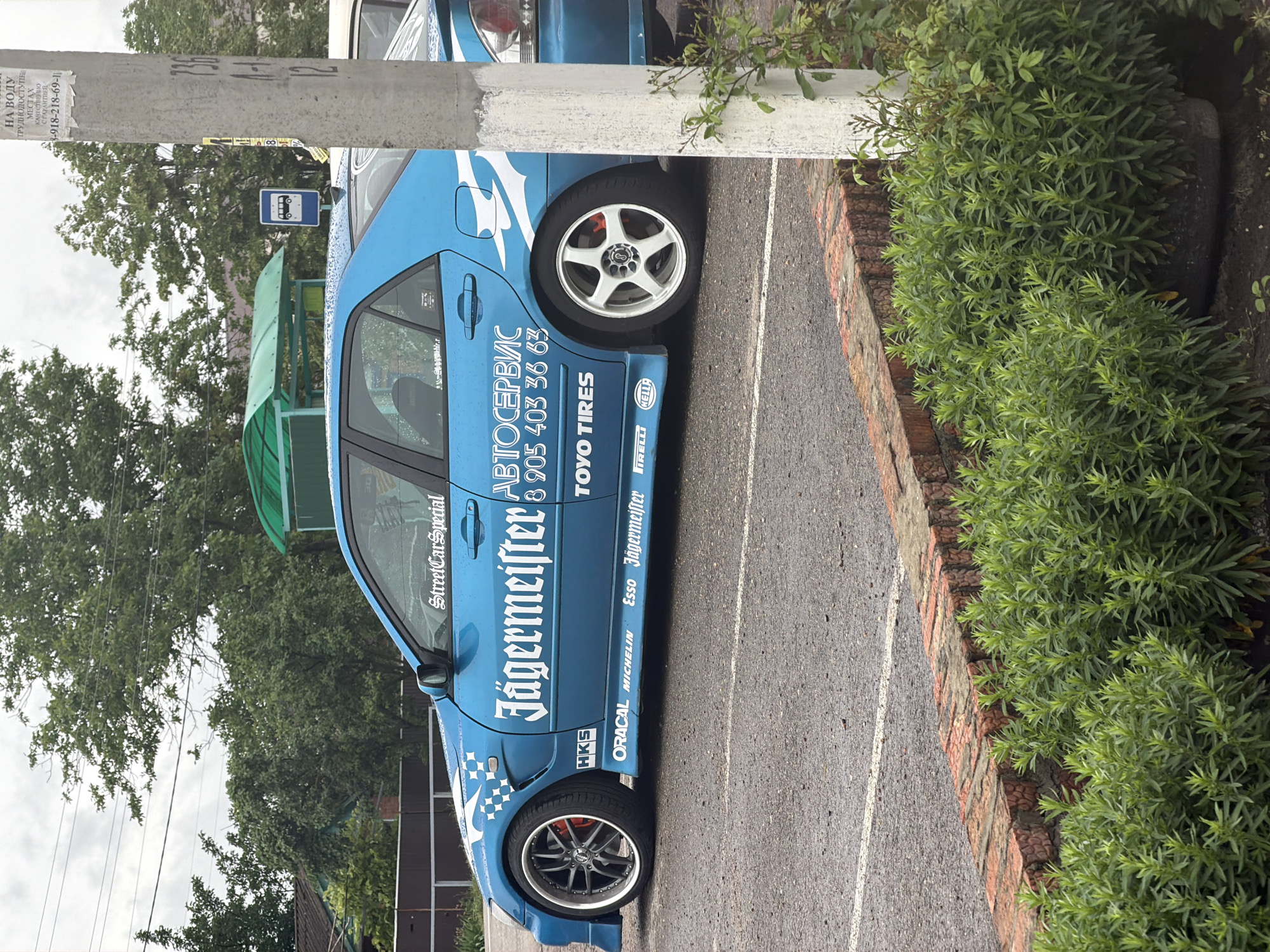 Here's a concise description of the scene:
A light blue car with racing decals and advertisements is parked on an asphalt parking lot or street. The car's decals prominently display "Jägermeister" and "Street Car Special." Other visible brands include Toyo Tires, Pirelli, Michelin, Esso, and HKS. Next to the car is a low brick border with shrubbery. In the background, there's a bus shelter and green trees. A concrete utility pole with a faded advertisement stands near the car. The location is Leningradskaya, Russia, based on the Russian Cyrillic script and phone number on the car.