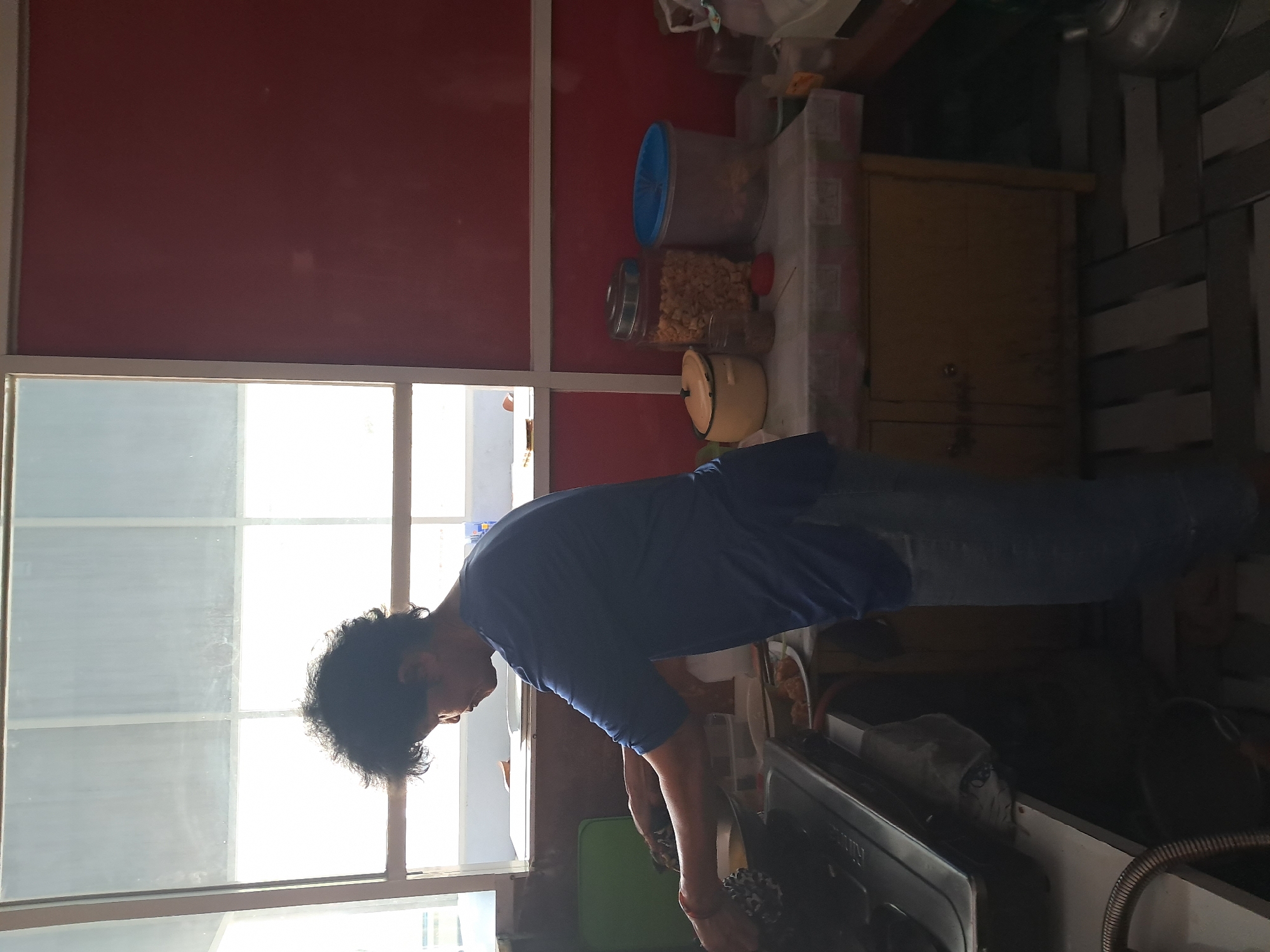 A person is standing in a kitchen, facing a gas stove, and appears to be cooking. The person is wearing a blue t-shirt and blue jeans. The kitchen has a red wall, a window with white blinds, a counter with various food containers, and a wooden cabinet. The floor is tiled with a striped pattern. The scene appears to be in a residential kitchen.