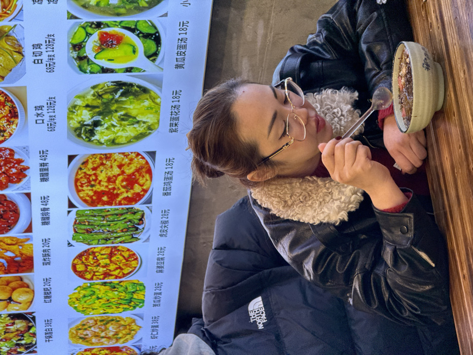 An indoor scene in a restaurant, likely within Yuzhong District, China, shows a woman eating from a bowl at a wooden table. The woman, positioned in the foreground and center-right, is seen in profile, leaning over a light-colored, patterned ceramic bowl. She is holding a metallic spoon in her right hand, scooping food from the bowl towards her mouth. Her hair is dark brown and appears to be pulled back. She wears gold-rimmed glasses, a dark leather-like jacket with a prominent cream-colored, curly fleece collar, and a reddish garment underneath. Dark nail polish is visible on her right thumb.
To the left and slightly behind the woman, a portion of a person wearing a dark blue puffer jacket with a white "THE NORTH FACE" logo on the left shoulder is visible, suggesting a second individual seated at the table. A grey hoodie is visible beneath the puffer jacket.
In the background, a brightly lit digital or printed menu board displays rows of various Chinese dishes with corresponding images and prices in Chinese Yuan. Visible dish names include "干锅蛋白 38元" (Dry Pot Egg White 38 Yuan), "红糖糍粑 20元" (Brown Sugar Glutinous Rice Cakes 20 Yuan), "现炸酥肉 20元" (Fried Crispy Pork 20 Yuan), "糖醋排骨 48元" (Sweet and Sour Ribs 48 Yuan), "口水鸡 68元/半支 128元/支" (Saliva Chicken 68 Yuan/half, 128 Yuan/whole), "麻婆豆腐 28元" (Mapo Tofu 28 Yuan), "番茄鸡蛋汤 18元" (Tomato Egg Drop Soup 18 Yuan), and "紫菜蛋花汤 18元" (Seaweed Egg Drop Soup 18 Yuan), among others. The food in the woman's bowl appears dark and granular, possibly a sweet or savory porridge-like dish with red and white specks. The table surface is made of light-brown wood with distinct horizontal grain patterns.