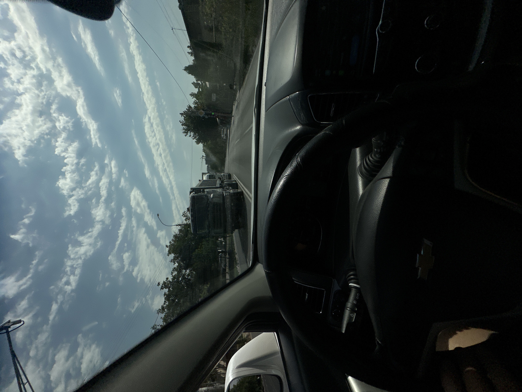 The image is a first-person perspective from inside a car in Tashkent, Uzbekistan. The driver's side of the car is visible, including the steering wheel with a Chevrolet logo. The road ahead shows traffic, with several trucks and other vehicles. Lush green trees line the side of the road, and the sky is partly cloudy with a bright sun. The scene appears to be a typical urban drive on a sunny day.