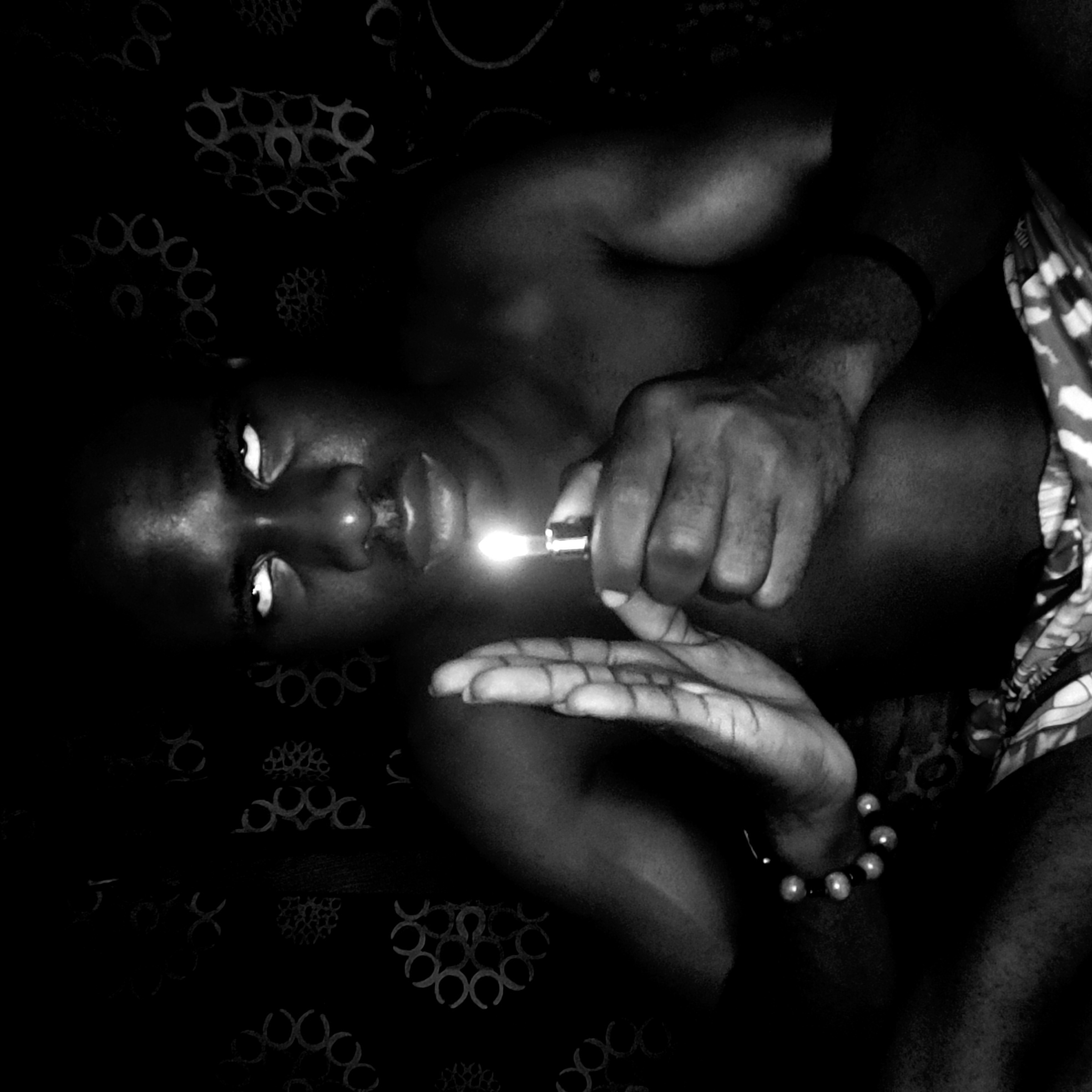 A black and white image of a person holding a lighter. The person is lying down on a patterned surface. The person's face is visible and their eyes are open and directed towards the camera. A lighter is held in one hand, and the flame is lit. The other hand is open, palm up. The person is wearing a bracelet on one wrist. The image appears to be taken in a dimly lit environment. There is no verifiable evidence of the location being Narayi, Nigeria.