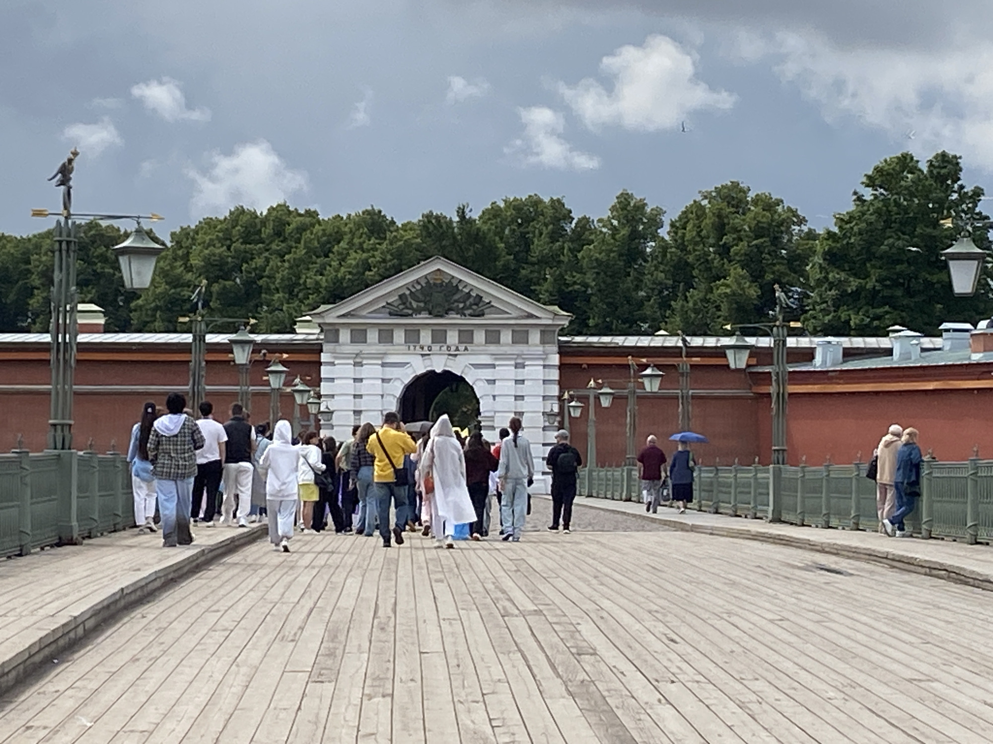 A group of tourists are walking on a wooden bridge towards an archway in a red brick building. The building has a white facade with an inscription that reads "1740 ГОДА". The sky is cloudy, and there are trees in the background. On the left side of the image, there are lampposts with decorative elements. The people on the bridge are dressed in casual clothing, and some are walking in pairs or small groups. The bridge is made of wooden planks, and there is a low green fence on either side. The scene appears to be of the Peter and Paul Fortress in Saint Petersburg, Russia.