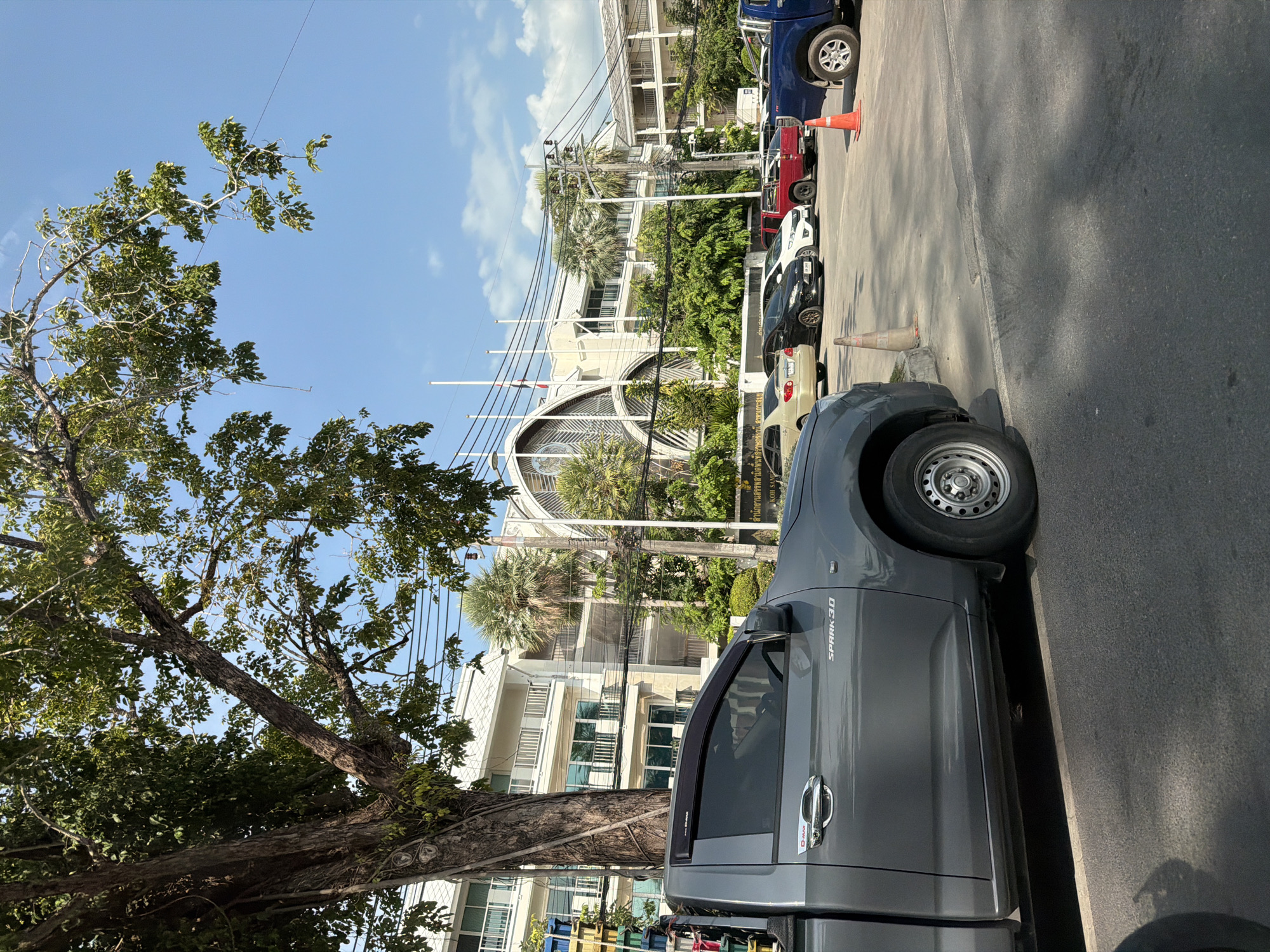 The image captures a street scene in the Ko Samui District of Thailand on a sunny day. In the foreground, the side of a grey pickup truck is visible, parked on the asphalt road. The truck has a chrome door handle and a "D-MAX SPARK 3.0" emblem on its side. To the left of the truck, a large tree with a thick trunk and leafy branches extends into the frame, partially obscuring a white building in the background. This building appears to be a multi-story structure with balconies and large windows. Beyond this building, more trees, including palm trees, are visible, along with power lines and utility poles. In the middle ground, several cars are parked in front of the building, including a gold sedan, a black sedan, a white hatchback, a red pickup truck, and a blue pickup truck. Traffic cones are placed on the road near the parked cars. The sky is clear and blue with a few wispy clouds. The overall impression is one of a typical urban street with parked vehicles and buildings, bathed in bright sunlight.