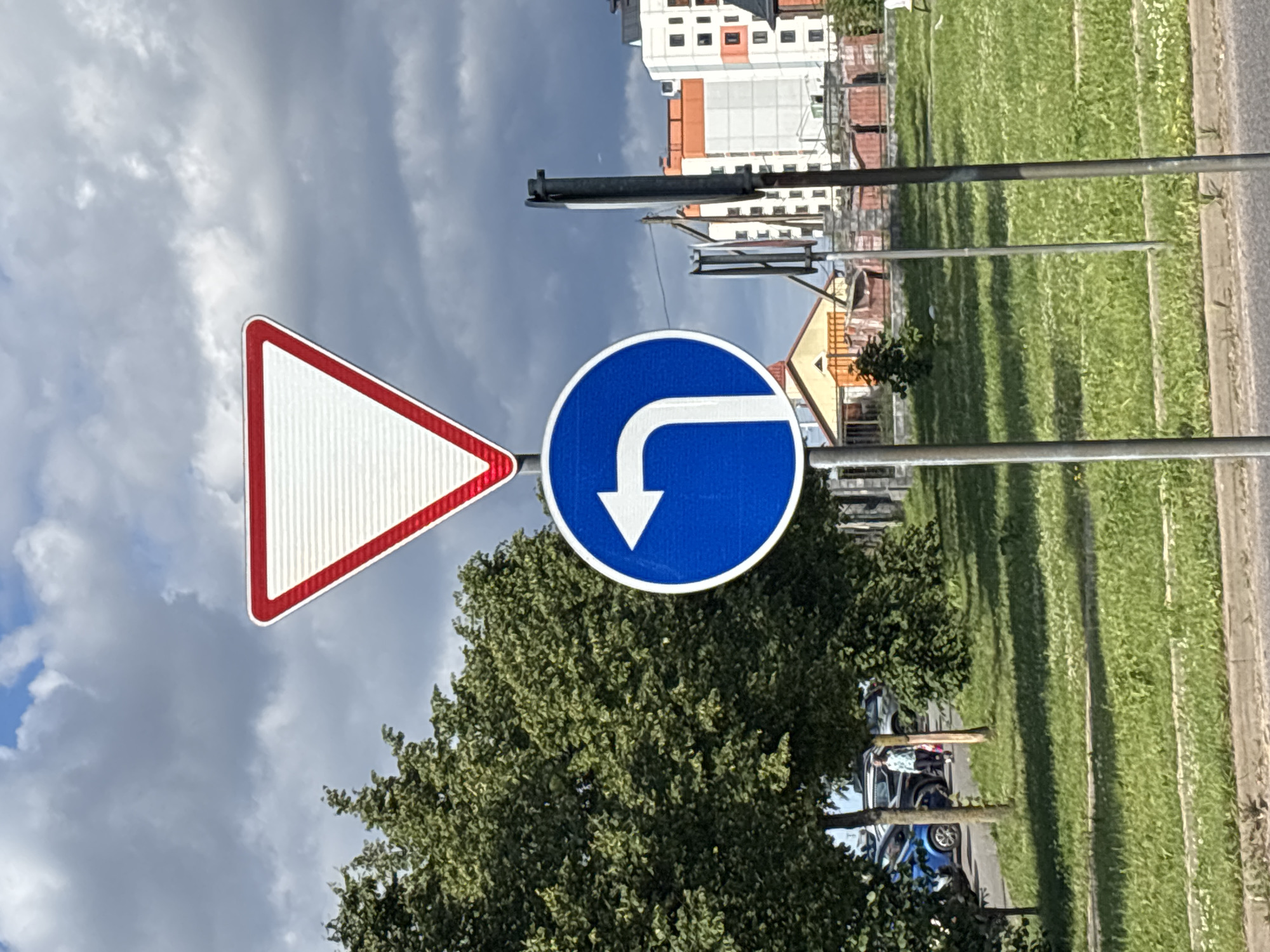 The image depicts a street scene with traffic signs and urban elements. A white triangular sign with a red border, commonly known as a "yield" or "give way" sign, is positioned at the top. Below it, a circular blue sign with a white arrow indicates a mandatory left turn. The signs are mounted on metal poles. In the background, there is lush green grass, trees, and modern buildings. Some cars are visible parked on a street in the distance. A person is walking in the middle ground. The sky is partly cloudy, suggesting it is daytime. The presence of a yield sign and a mandatory left turn sign, along with the characteristic architecture of the buildings, suggests this location could be in Minsk, Belarus.