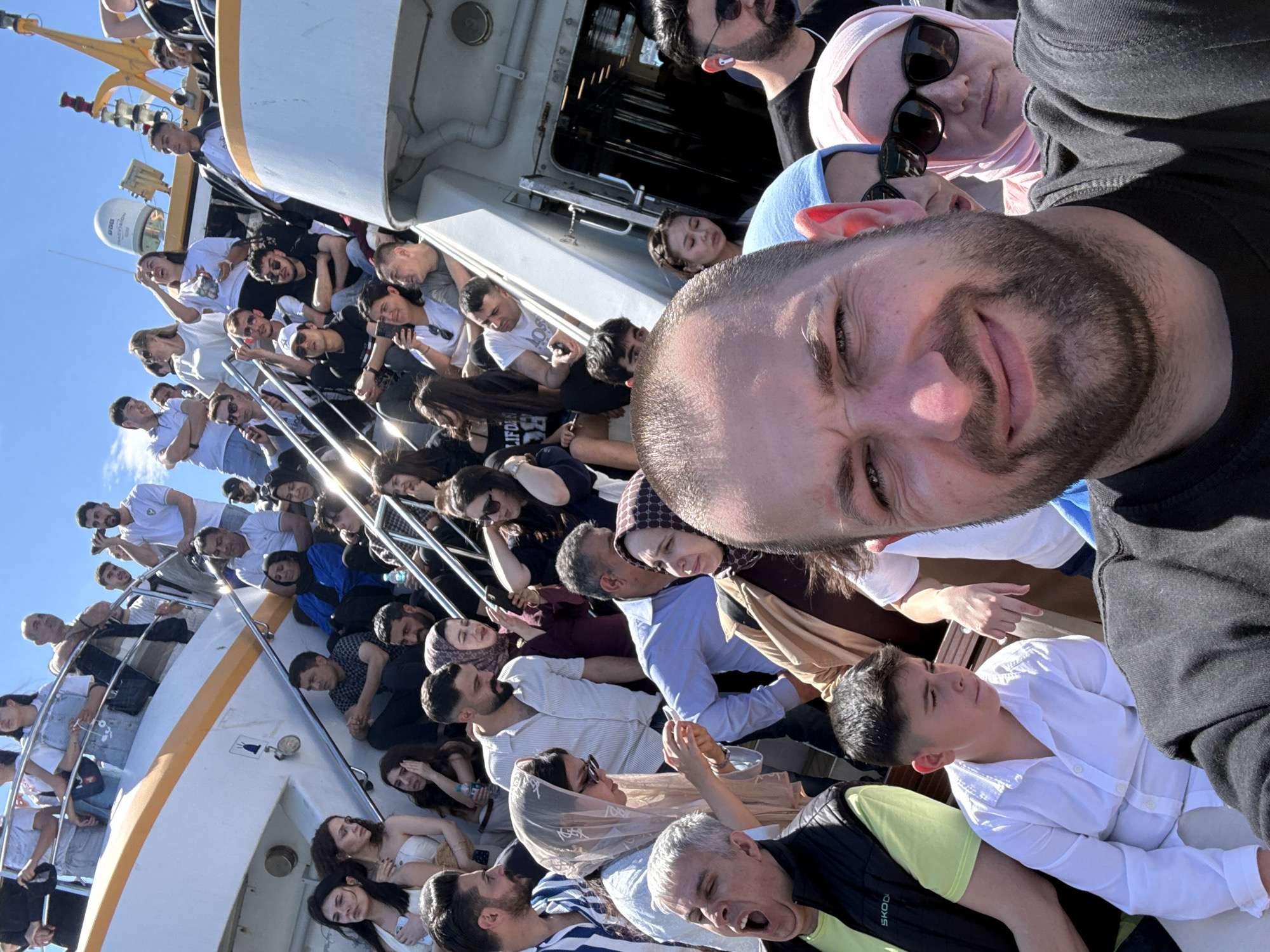 The image shows a large group of people on a ferry in Kadıköy, Turkey. The view is from the perspective of someone taking a selfie. The selfie includes the person holding the camera, as well as two people behind them, one with sunglasses and a headscarf, and another with a blue headscarf. The rest of the ferry is visible, with numerous people standing along the railings or sitting inside the ferry. The sky is clear and blue. The ferry has white and yellow paint.