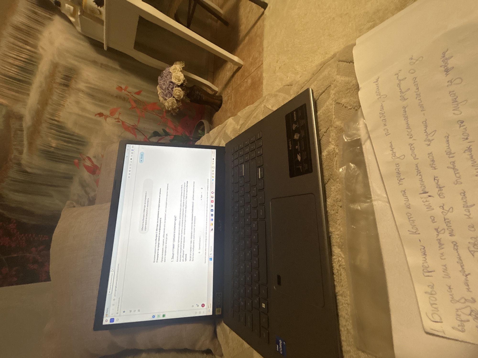 The image shows a laptop displaying a web page with information about error correction in computers. Next to the laptop is a piece of paper with handwritten notes in Bulgarian that also discuss bit errors and data transmission over WiFi. The laptop is an Acer Aspire 5, with an Intel Core i7 processor. The scene is likely in a domestic setting, possibly a home in Burgas, Bulgaria, given the Bulgarian text and the appearance of the surroundings. There is a couch with a waterfall picture behind the notebook. There is also a decoration with white roses on a dresser.