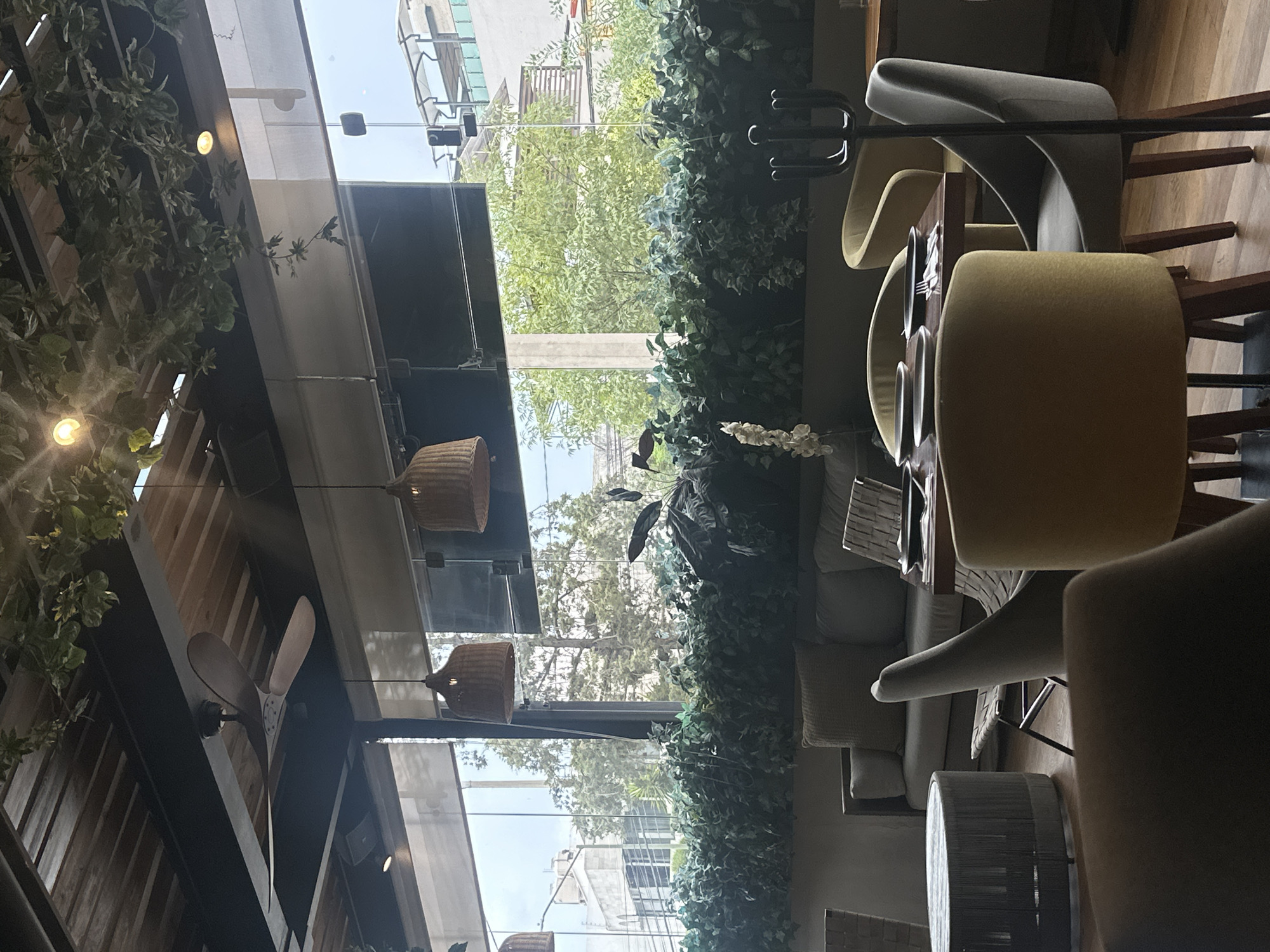 The image depicts the interior of a restaurant in Huixquilucan de Degollado, Mexico. The restaurant features a combination of indoor and outdoor elements, with large windows offering a view of greenery and buildings outside. The interior design incorporates natural materials, such as wooden beams and woven lampshades, along with decorative plants and foliage. The furniture consists of upholstered chairs and sofas, along with dining tables set with plates and cutlery. A ceiling fan is visible in the upper left corner of the frame. The overall atmosphere is modern and inviting, with a focus on natural light and greenery.