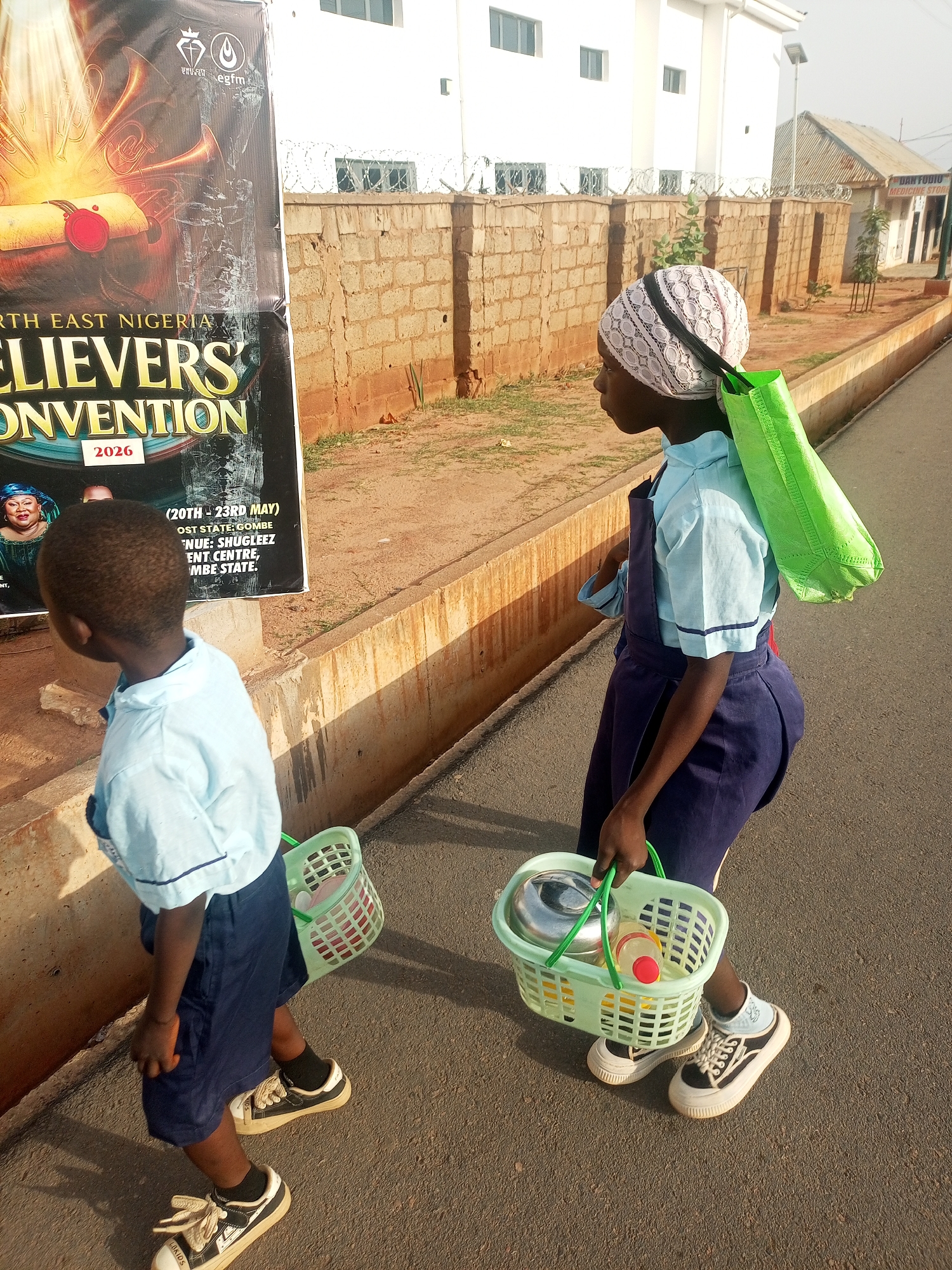 The image shows two children, likely students, walking on a street in what appears to be an urban area. A young boy is on the left, wearing a light blue shirt and dark blue shorts, and carrying a small green plastic basket. A slightly older girl is on the right, dressed in a similar uniform, but with a darker blue overall-style dress over her shirt, and a head covering. She carries a green shopping bag over her shoulder and a green basket filled with what appears to be food containers. In the background is a wall topped with barbed wire, a building, and a sign promoting a "Believers' Convention 2026" event, which is located at "Shugleez Entertainment Centre, Gombe State." A sign for "Dan Fodio Medicine Store" is visible in the far background. The location appears to be in Nigeria.
