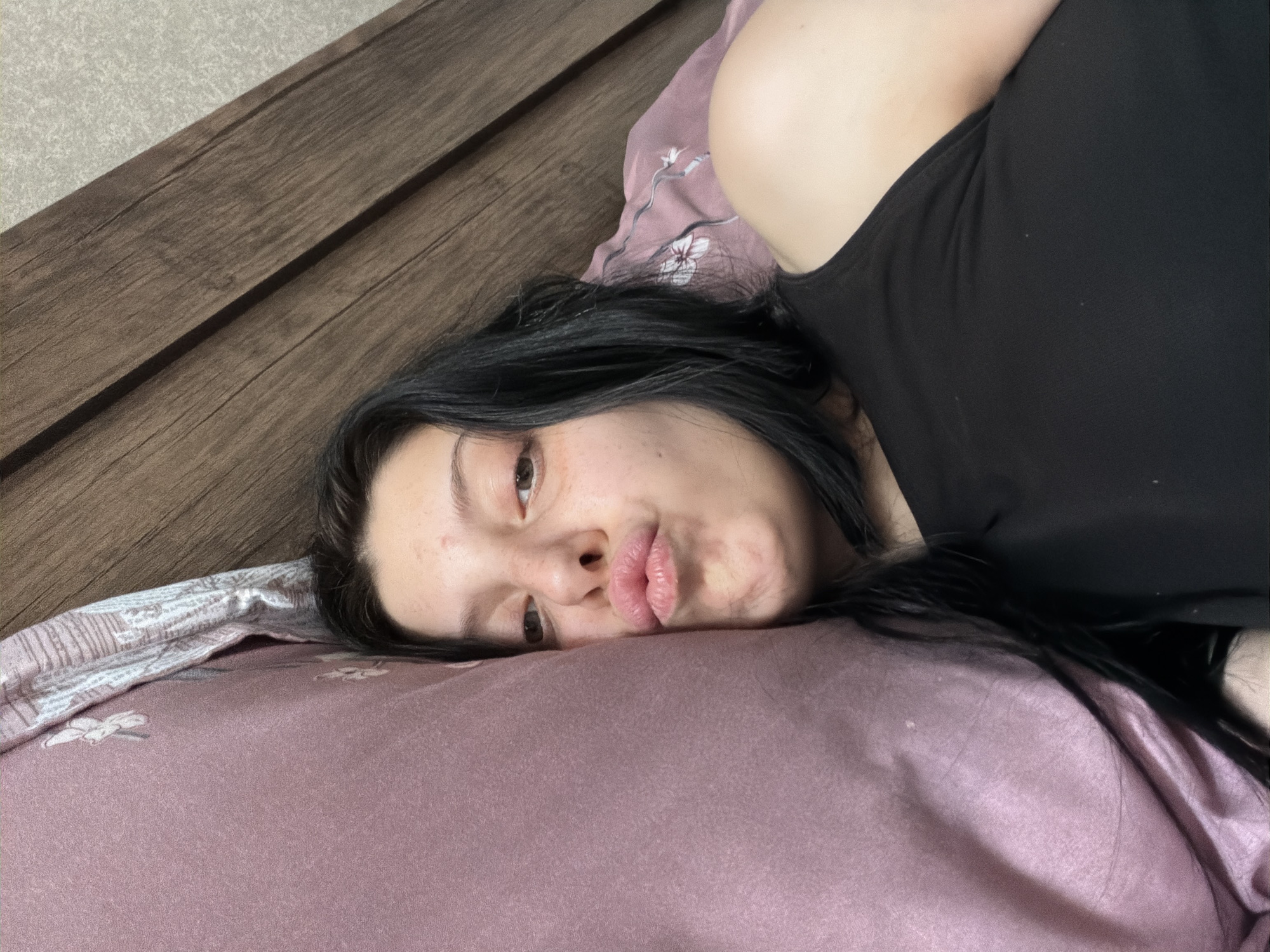 A young woman with dark hair and light skin is lying in bed, facing the camera. She is wearing a black tank top and has her lips puckered as if for a kiss. Her head is resting on a pink pillow. The background shows a wooden headboard and a grey textured wall. The scene is indoor and appears to be a bedroom. There is no visible indication of the location being in Tbilisi, Georgia.