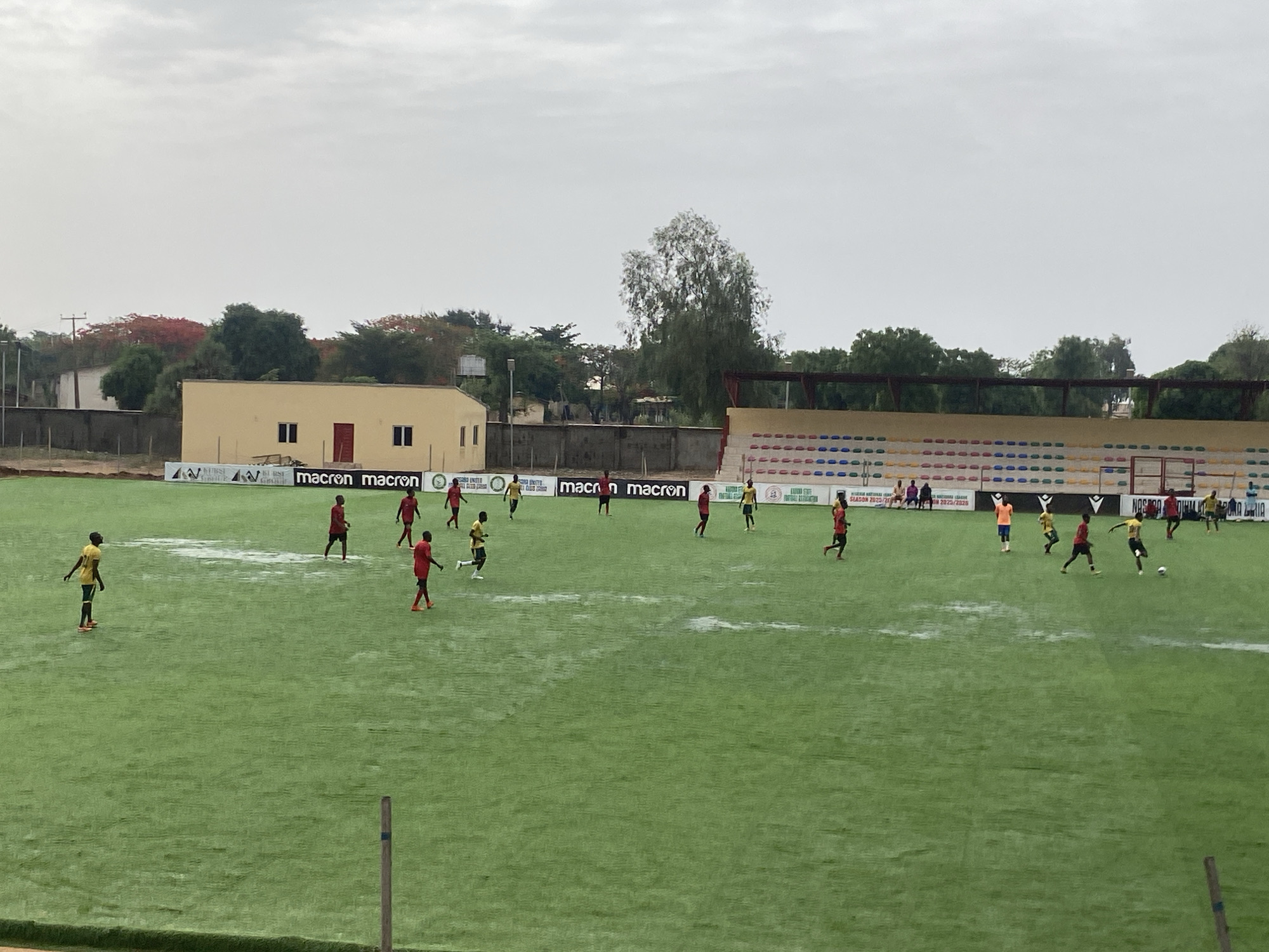 The image depicts a soccer match in progress at a stadium located in Tudun Wada, Nigeria. Two teams, one in red uniforms and the other in yellow and black, are actively engaged in play on a green artificial turf field. The field shows signs of recent rainfall, with visible puddles in multiple locations. The stands are visible behind the field, only partially filled with spectators. A building with a tan exterior and red door is visible behind one of the field's sidelines. The sky is overcast, suggesting inclement weather. Signage with the "macron" brand is displayed along the sideline. Trees surround the outer perimeter of the stadium, obscuring a clear view of the surrounding cityscape.