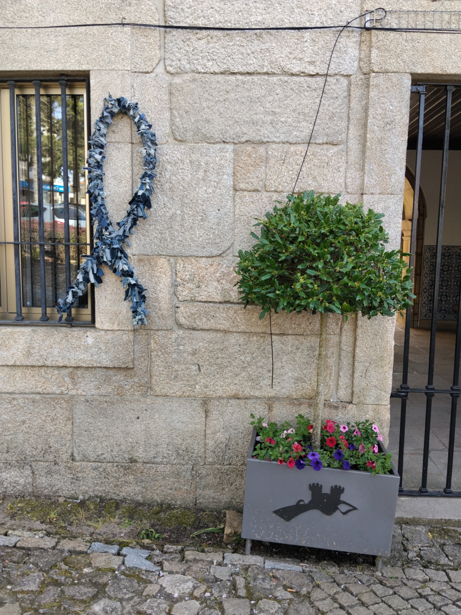 The image shows a stone wall in Penedono, Portugal. A window with iron bars is on the left. A blue denim ribbon hangs beside it. To the right, a topiary tree in a gray planter is positioned next to a doorway with an iron gate. The planter features a silhouetted emblem of a castle and a bird. Cobblestones cover the ground.