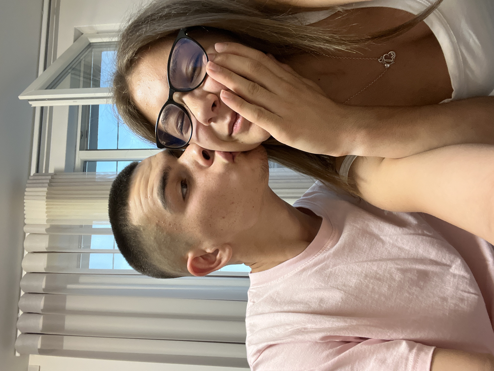 An indoor, eye-level selfie captures two individuals, a man and a woman, in an affectionate pose within a residence in Karavelovo, Bulgaria. The man, positioned on the left, has short dark hair with faded sides, and is wearing a light pink crew-neck t-shirt. He is seen leaning in to kiss the woman's right cheek. The woman, on the right, has light brown hair and is wearing black-framed eyeglasses. Her eyes are closed, and her right hand is gently touching her chin/cheek. She is wearing a white tank top, revealing a silver necklace with a heart-shaped pendant inscribed "love" and a small round charm. In the background, white pleated curtains are visible on the left, and a white-framed window is partially open on the right, showing a clear blue sky outside.