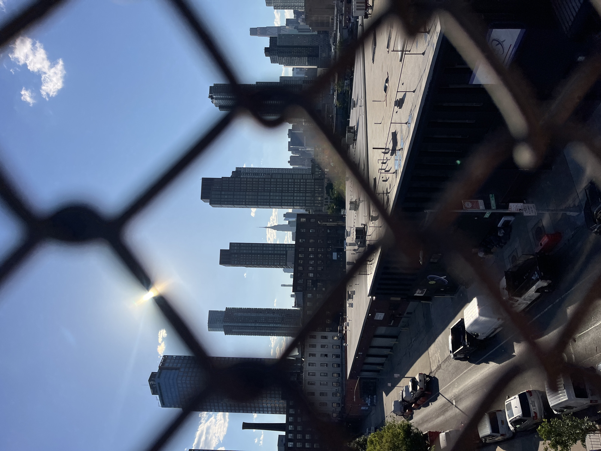 The image is a vertical, eye-level, medium shot of an urban landscape viewed through a chain-link fence. The sun is prominently visible in the upper center of the frame, casting lens flare and rays into the clear blue sky, which is dotted with scattered white clouds. In the mid-ground, a row of industrial-style buildings stretches horizontally across the frame, with flat rooftops featuring HVAC units and other rooftop equipment. Some of these buildings appear to be warehouses or factories.
In the foreground, a street runs parallel to the buildings, with several vehicles parked and driving along it. These vehicles include cars, vans, and pickup trucks, some of which appear to be commercial vehicles. A person can be seen on the sidewalk in the lower right portion of the frame, but they are indistinct due to the fence's blurriness.
The background reveals a dense cityscape dominated by numerous skyscrapers of varying heights and architectural styles, indicative of a major metropolitan area. The iconic Empire State Building is identifiable among the distant skyline. The overall scene suggests a view from an elevated position overlooking a commercial or industrial district within New York City, possibly Queens or Brooklyn, given the proximity to the Manhattan skyline. The time of day appears to be late afternoon or early morning due to the angle of the sun and the shadows cast on the street.