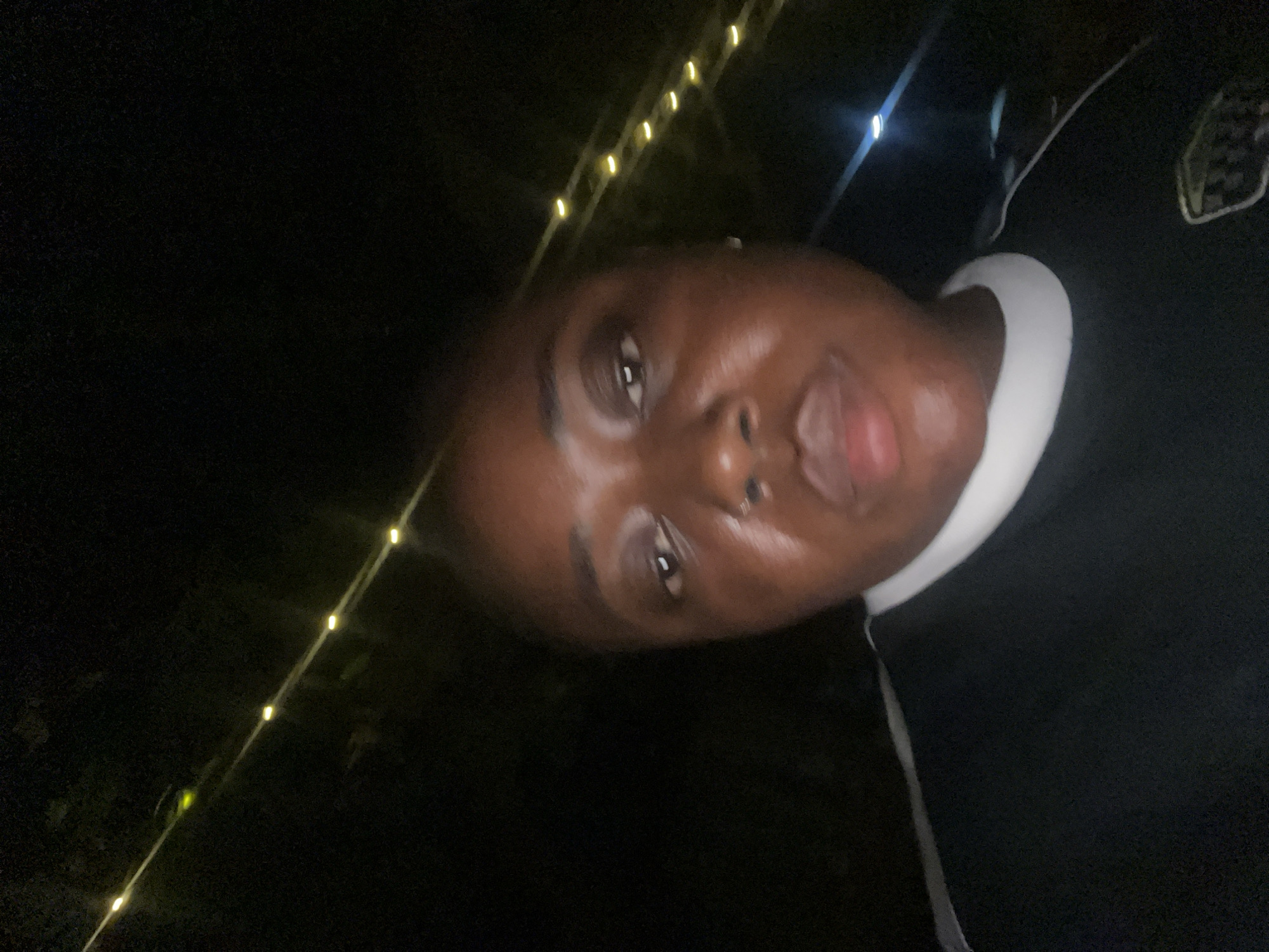 This nighttime portrait, taken in Abuja, Nigeria, features a single individual, an adult female with dark skin, looking directly at the camera. Her face is centered, exhibiting a neutral expression, and she has a silver-toned ring piercing in her left nostril. Her skin shows some reflectivity, possibly due to natural oils or sweat. She wears a dark-colored top with a prominent white collar and white trim detail along the shoulders. A faint, possibly checkered or striped, patch is visible on the lower right side of her garment. The background is largely dark, illuminated by a diagonal string of warm-toned decorative lights stretching across the upper portion of the frame. A brighter, blue-toned light source is also visible in the distant background on the right.
