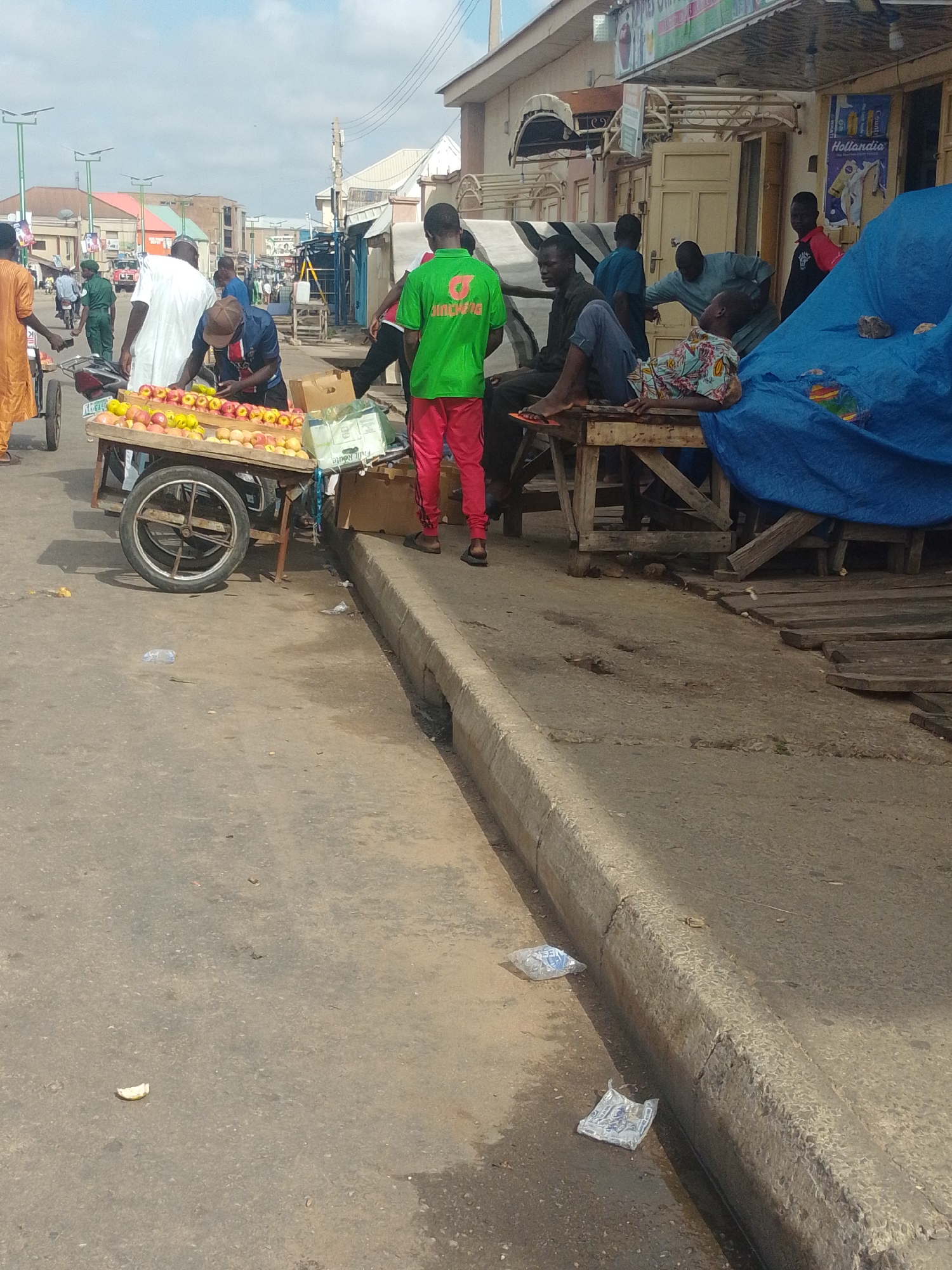 The image depicts a street scene in Pantame, Nigeria, likely on a market day. In the foreground, a fruit cart is laden with apples. Several individuals are present: a person in an orange garment stands near the cart, possibly a customer or vendor. Another person, wearing a green t-shirt with a logo and red trousers, stands facing away from the camera, appearing to interact with the fruit. A man in a white robe is also visible, standing behind the fruit cart. Further back, other people are seen around stalls and buildings lining the street, some seated on wooden structures. The environment is urban, with a paved road, a concrete curb, and various buildings and streetlights visible. There is evidence of litter on the ground, including plastic bags and wrappers, suggesting a busy public space. The sky is overcast.