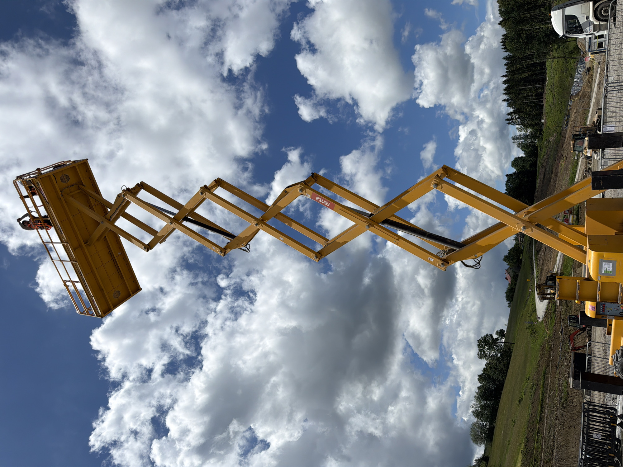 The image depicts a tall, yellow scissor lift in the foreground, extending upwards against a blue sky with fluffy white clouds. Two construction workers, wearing orange safety vests, are visible on the elevated platform of the scissor lift. The scissor lift itself is a complex mechanical structure with interconnected metal beams forming a crisscross pattern. In the background, a green, hilly landscape is visible, with trees dotting the slopes and a winding road leading upwards. Further in the distance, a dense forest stands at the top of the hill. A white truck is parked to the right of the scissor lift. The overall scene suggests a construction site in a rural or semi-rural area. While the prompt mentions Janowice, Poland, there is no explicit visual evidence within the image to confirm this specific location. The environment appears to be an outdoor setting, possibly undergoing development or construction, given the presence of heavy machinery and the unfinished appearance of some areas in the mid-ground.