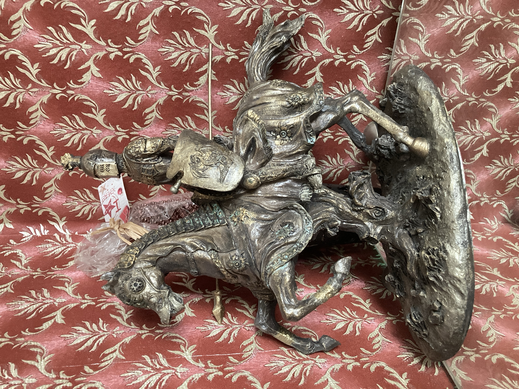 The image shows a detailed bronze statue of a knight on horseback. The knight is wearing armor, a helmet, and is holding a shield and a lance. The horse is depicted in mid-gallop, with its front legs raised. The statue is placed on a textured base that resembles rough terrain. Behind the statue is a red fabric with a gold damask pattern. There is also a tag attached to the knight's head with some writing on it and what appears to be a bag of small beads. The lighting suggests it is indoors, and the statue is resting on a glass surface. The overall scene is a close-up shot of the statue against its decorative backdrop.